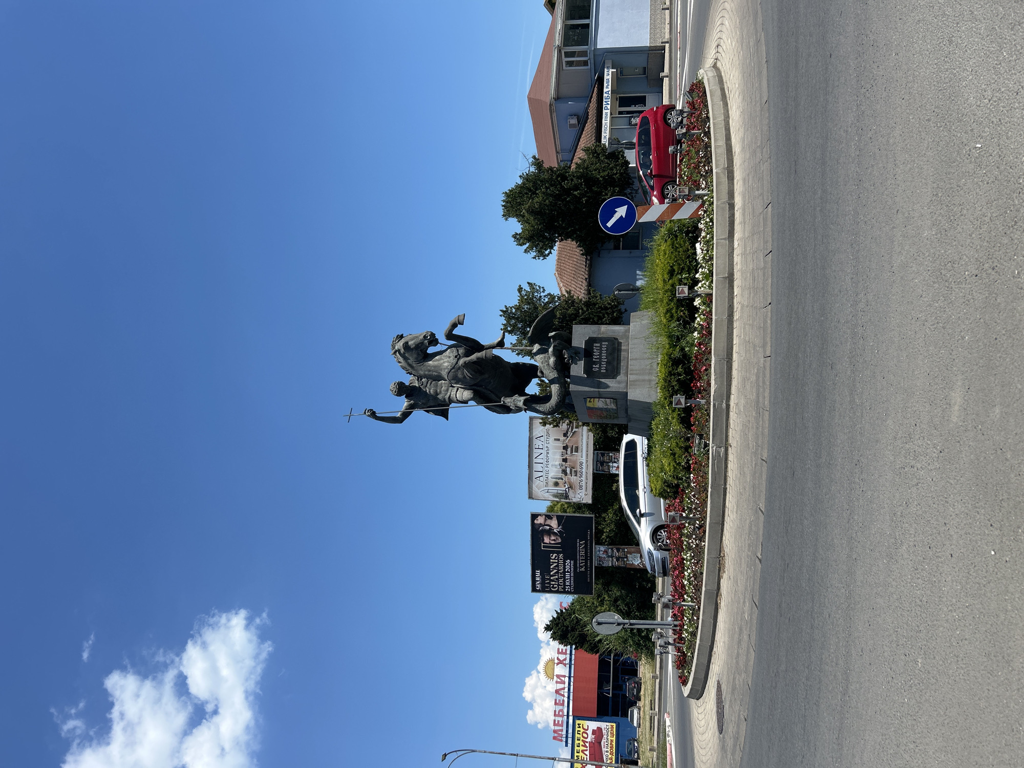 This is a full-shot photograph of a roundabout in the city of Pomorie, Bulgaria, on a clear, sunny day with a bright blue sky and scattered white clouds. Dominating the center of the roundabout is a large bronze equestrian statue of Saint George slaying a dragon. The statue stands on a pedestal with Cyrillic inscriptions. The roundabout is adorned with a circular flower bed filled with red and white flowers.
Surrounding the roundabout, several elements of the urban environment are visible. To the left, a large billboard features advertisements for "MEBELI XES" (furniture) and "GINNIBALL LIVE GIANNIS XARTARNIOS." Further left, a street lamp is visible. In the background, beyond the roundabout, are several buildings, including a light blue structure with signage that includes "MALIK FISCH FISH РИБА РЫБА ШАБ." A red hatchback car is driving around the roundabout, and a silver SUV is parked in the background near one of the billboards. Road signage, including a blue circular sign with a white arrow pointing left, is also present. The road surface of the roundabout and the surrounding streets are visible.