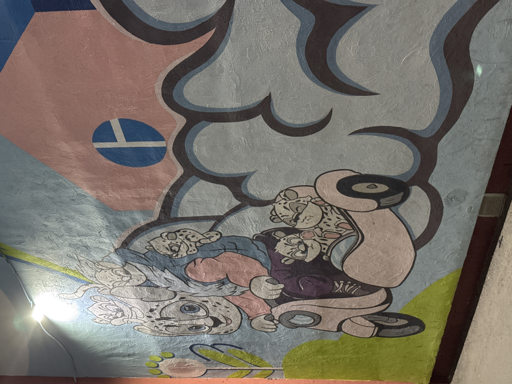 This is a mural or painted artwork. It depicts several cartoonish characters resembling snow leopards. They are in a pile or grouping, with one character appearing to be upside down. The characters are wearing clothing, including what appears to be a hooded sweatshirt, shoes and pants. They are set against a blue, pink, and green background with stylized cloud-like shapes outlined in black. The background also includes a symbol of a plus sign inside of a circle. The artwork appears to be on an interior wall, possibly a ceiling. The photograph was taken under artificial light. It is located in Kazan, Russia.