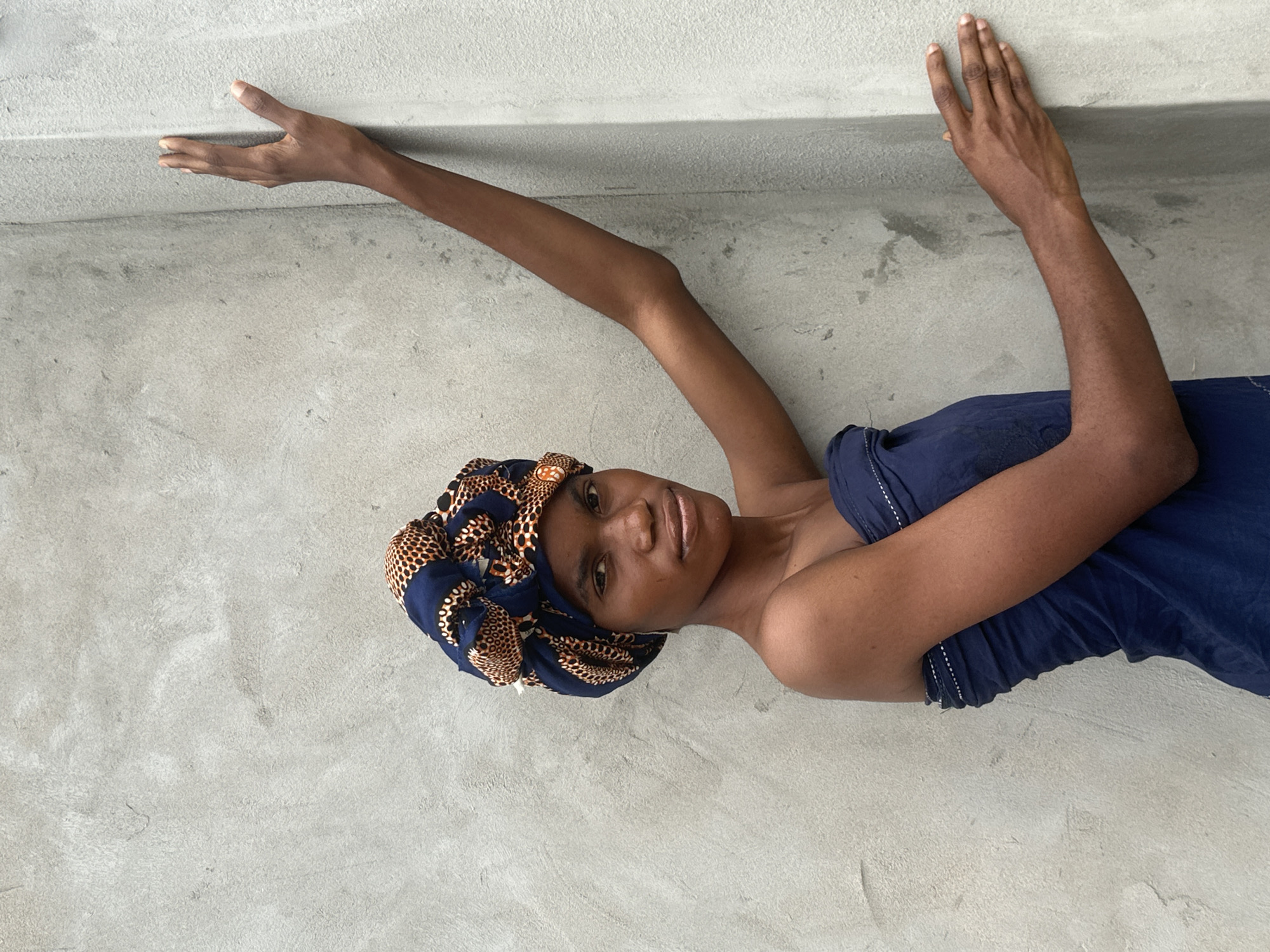 A young Black woman stands against a concrete wall in Badagry, Nigeria. She is wearing a dark blue strapless dress and a patterned headwrap with dark blue, orange, and black colors. Her right arm is extended upward, with her hand resting on the wall, while her left arm is bent, with her hand near her waist. The wall appears to be unfinished, with a rough texture. The lighting suggests it is daytime.