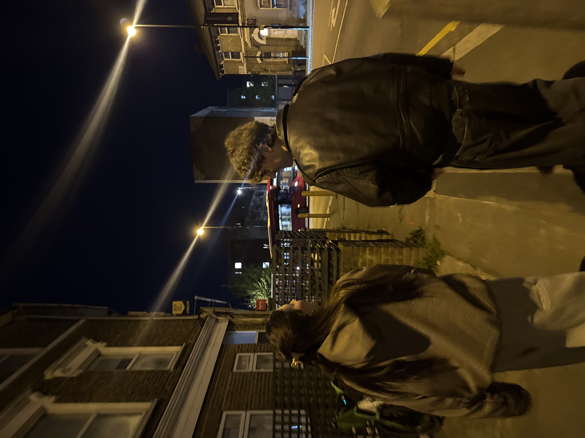 Two individuals are standing on a sidewalk near a street at night. The first individual, presumably male, is wearing a black leather jacket. The second individual, seemingly female, is wearing a dark-colored coat or jacket and light-colored trousers. Streetlights illuminate the scene. Buildings line the street. A red double-decker bus is visible in the background. The location is likely in London, United Kingdom.