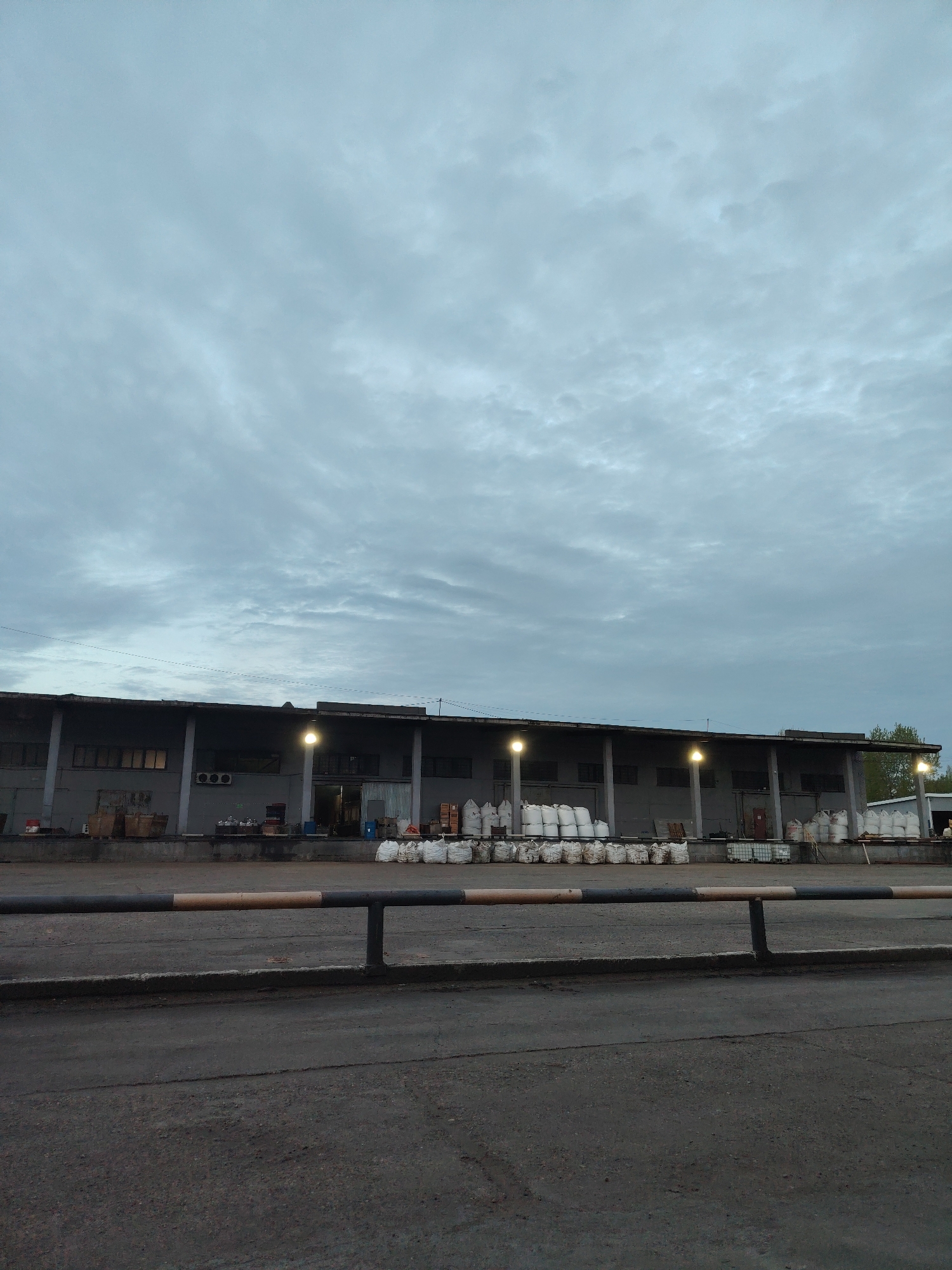 The image depicts a low-angle view of a one-story industrial building in Noviy, Russia, under an overcast sky. The building has a series of loading bays with support pillars and evenly spaced lights illuminating the loading area. White sacks are stacked along the loading dock. In the foreground, a barrier of black and tan horizontal poles is present. The ground in front of the barrier is paved and shows signs of wear and use. No people are visible in the image.