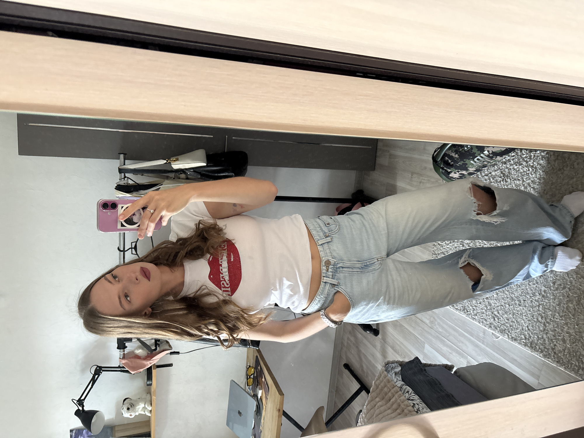 A young woman with long, wavy brown hair is taking a mirror selfie in what appears to be a bedroom or dressing area. She is wearing a white t-shirt with a red lip graphic and the text "Lipstick" on it, and light-wash, ripped jeans. Her left arm is raised, holding a pink phone to take the picture. Her right hand is resting on her hip.
The background is a mirror reflecting the room. Visible in the reflection are a closet with some clothes and bags hanging, a desk with a laptop, a lamp, and a stuffed animal. The overall lighting suggests it is daytime. The setting appears to be indoors, possibly an apartment in Perm, Russia. The woman's expression is neutral, and she is looking directly at her phone's camera.