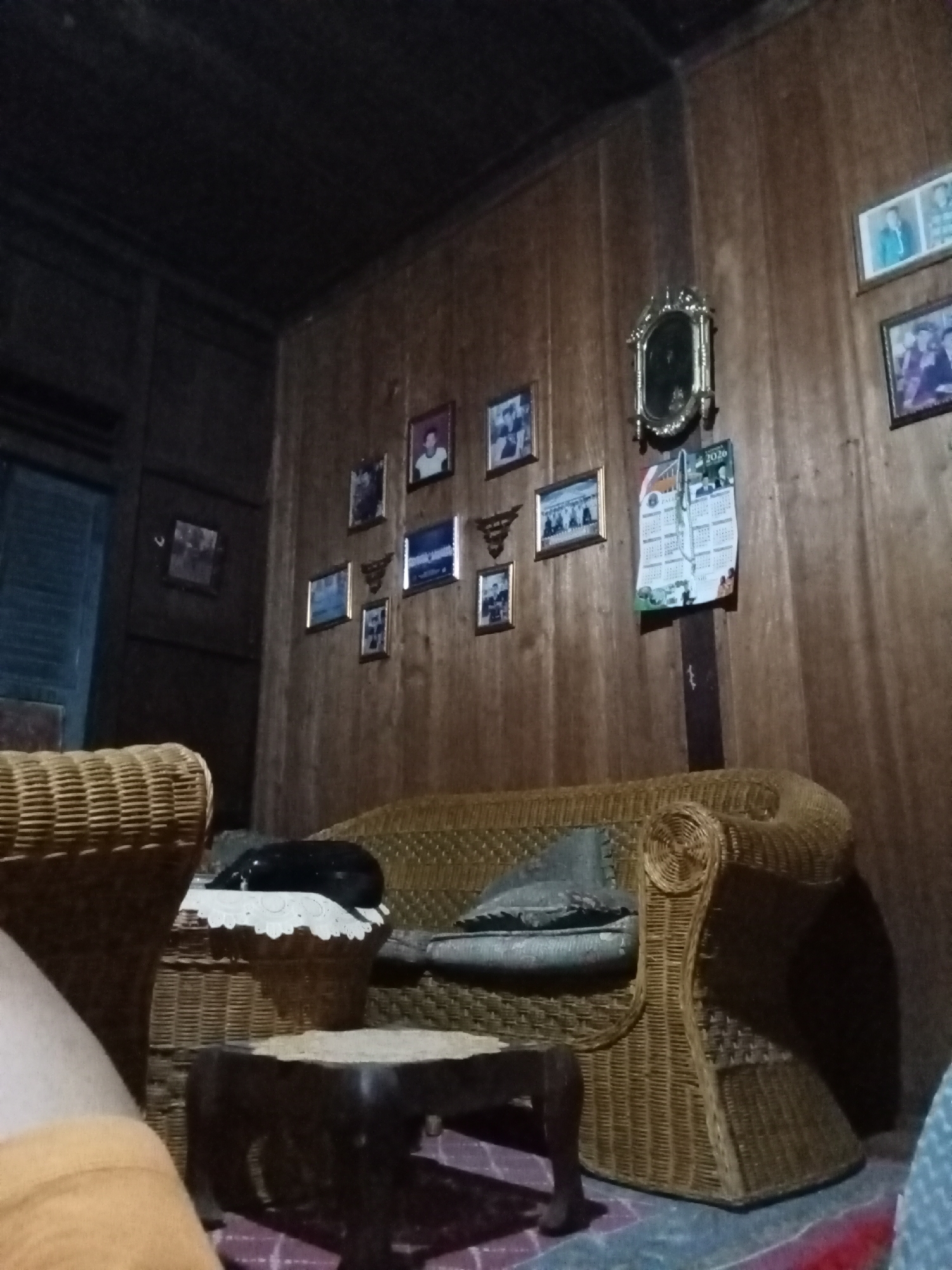 The image depicts a dimly lit living room, likely in Setia Jaya, Indonesia. The walls are paneled with dark wood, and several framed photographs and a decorative mirror adorn them. In the foreground, a woven wicker sofa with cushions is partially visible. To the left, another woven wicker chair is present. A small, dark wooden table with carved legs sits on a patterned rug in front of the sofa. A black object, possibly a pet, rests on a white doily atop a woven basket on the sofa. A calendar from 2020 is hanging on the wall. The overall atmosphere is domestic and somewhat dated.