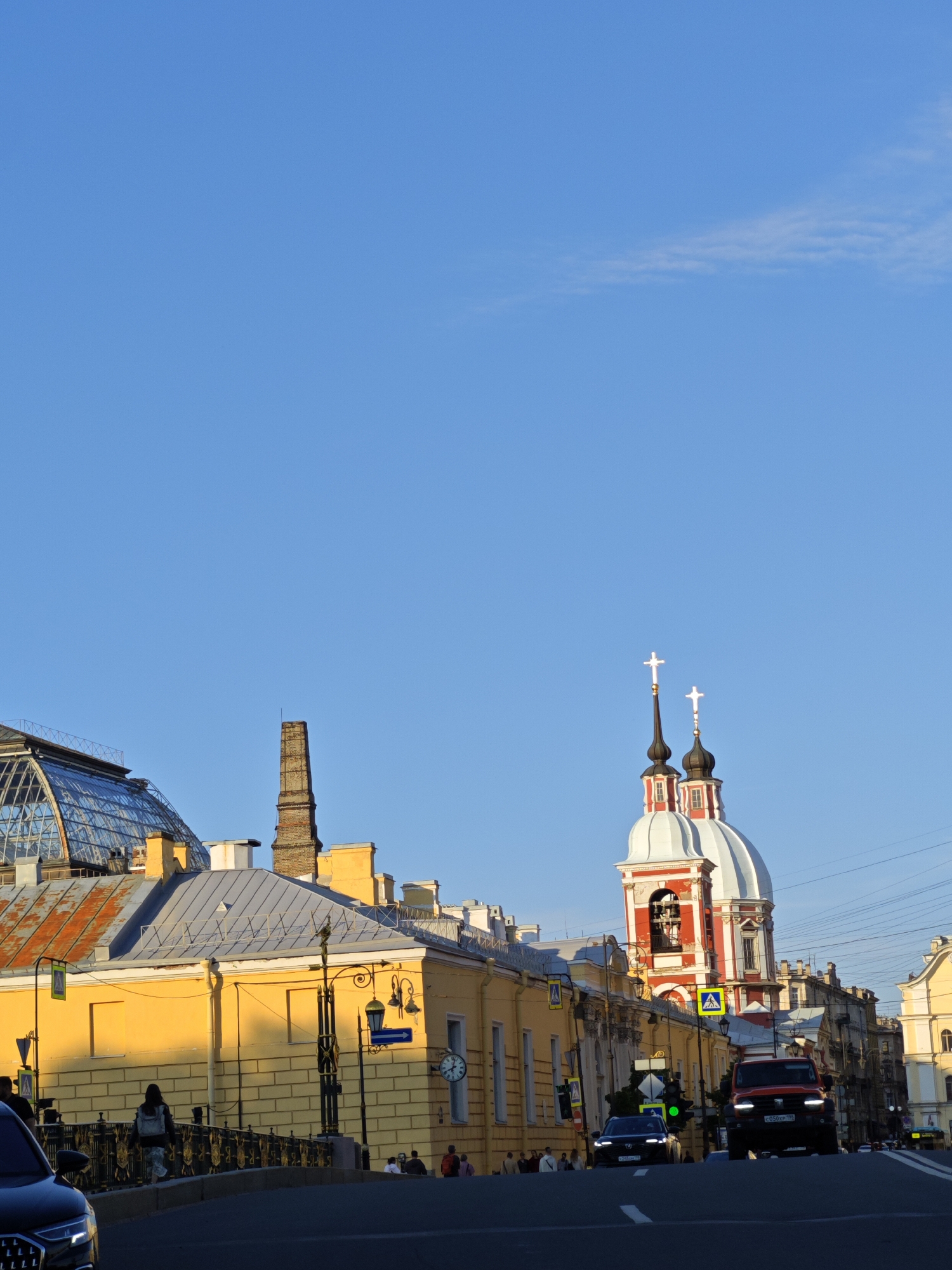 This image depicts a street scene in Saint Petersburg, Russia, likely in the vicinity of the historic city center, given the presence of classical architecture and a distinct church dome.
In the foreground, a dark-colored car is partially visible on the left, facing forward. Beyond it, a traffic light shows green. The road is paved and appears to be a multi-lane street. On the right side of the road, a red SUV is driving forward. A clear blue sky with a faint wisp of cloud stretches across the upper half of the image.
Dominating the mid-ground is a yellow building with a gray, gabled roof. A distinctive brick chimney stands tall to the left of this building. To the right of the yellow building, a domed church with red and white accents and two crosses on its spires is prominent. The church features arched windows and a bell tower. A clock is visible on the yellow building.
A pedestrian bridge with ornate railings is situated in front of the yellow building, with a few people walking on it. A woman with a backpack is visible on the bridge. Streetlights and traffic signs are present along the street and on the bridge. The buildings on the right side of the street are a mix of architectural styles, suggesting a blend of historical and perhaps more modern construction. The overall lighting suggests it is either early morning or late afternoon, with warm sunlight illuminating the facades of the buildings.