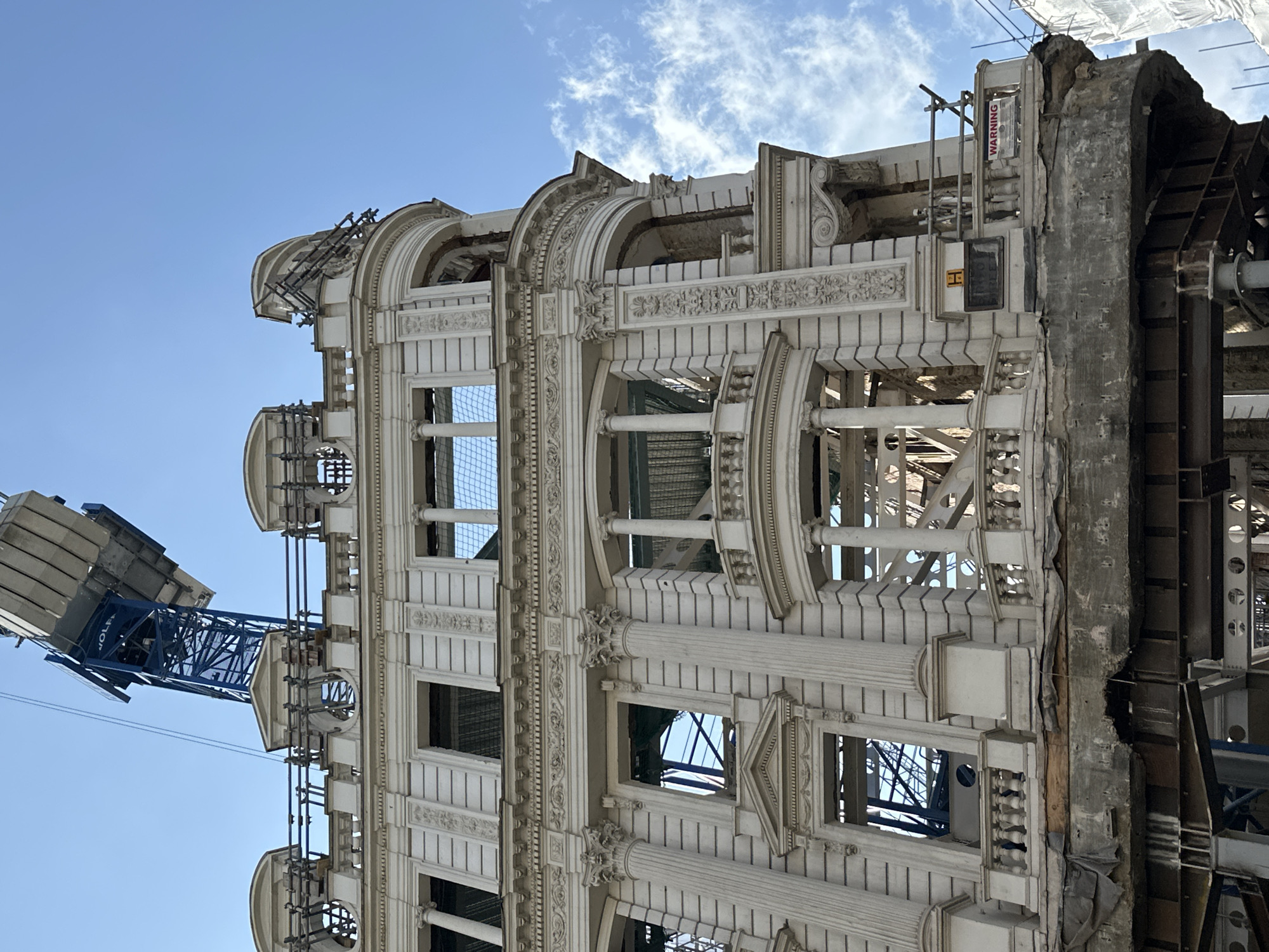 The image shows a ornate, light-colored building facade undergoing deconstruction or significant renovation, with only the exterior shell preserved. The architectural style features classical elements, including arched window openings, decorative pilasters with capitals, intricate friezes, scrollwork, and balustrade details. Most windows are empty openings, revealing the exposed internal metal framework, scaffolding, and safety netting within.
On the far right, the facade ends abruptly, exposing raw concrete and heavy steel support structures that buttress the remaining historical wall. A red sign with the word "WARNING" is affixed to scaffolding on this exposed section. Below it, a partially visible white sign displays "BAY TREE" and hints of "STREET."
In the background on the left, a large blue tower crane, partially visible with the name "WOLFA" on its arm, extends against a clear blue sky with a few wispy white clouds. The bright, even lighting indicates a sunny daytime scene. The image captures an urban landscape in transformation, showcasing the contrast between the preserved historic facade and the active construction or demolition work behind it.