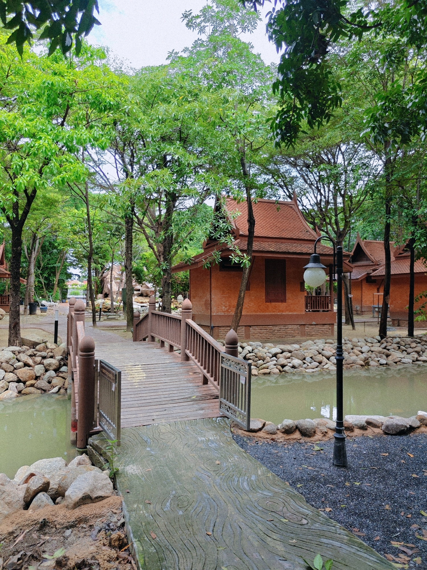 The image depicts a serene outdoor scene, likely within a resort or cultural park, in Hantra Subdistrict, Thailand. A wooden bridge with railings arches over a murky, calm body of water. The bridge leads from a textured concrete pathway with a wood-grain pattern to a gravel path.
On the far side of the water, traditional Thai-style buildings with terracotta roofs and orange-brown walls are visible, nestled amongst lush green trees. The architecture suggests a cultural or historical theme. The water is bordered by a collection of large, grey stones.
A black lamppost with a globe-shaped light fixture stands near the water's edge, to the right of the bridge. The ground around the buildings and pathways is covered in a mixture of gravel, sand, and fallen leaves. The overall impression is one of tranquility and natural beauty combined with architectural interest. There are no people visible in the image.