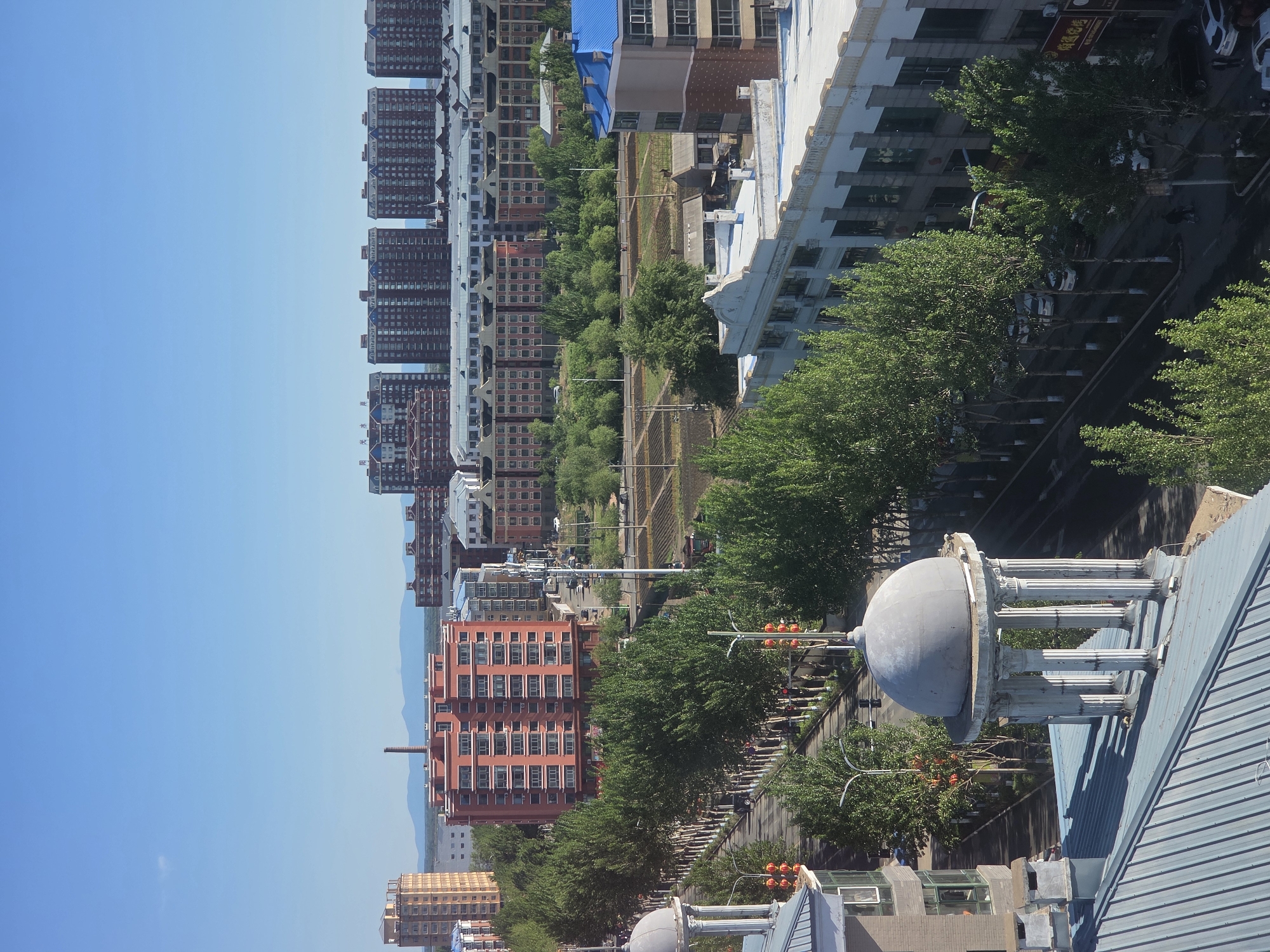This image presents an expansive, sunlit urban landscape of Tongjiang, China, captured from an elevated vantage point. A wide, clear blue sky dominates the top portion of the frame, meeting the faint outlines of distant mountains on the horizon.
The city sprawls below, characterized by a mix of multi-story buildings. On the left side of the frame, numerous residential or commercial high-rises with dark red or brown facades and many windows stand in rows. One of these buildings clearly displays the Chinese characters "阳光水郡" (Yángguāng Shuǐjùn), which translates to "Sunshine Water County" or "Sunshine Waterfront." On the right side, other buildings with lighter exteriors and some blue roofs are visible.
A prominent feature cutting horizontally through the middle ground is a wide, tree-lined green belt or parkway, providing a natural divide within the urban environment. Roads, streetlights, and traffic signals are visible, with several vehicles, including parked cars, indicating ongoing urban activity. In the bottom right corner, a distinctive architectural element—a grey dome supported by columns, possibly a cupola or gazebo—is seen on a rooftop, belonging to a building closer to the viewer. Another faint brown sign on a building to the right includes Chinese characters like "商贸" (shāngmào), suggesting commercial establishments.
The bright, clear light and distinct shadows indicate a sunny day, likely midday or early afternoon. The overall impression is one of a well-developed and verdant city under pleasant weather conditions.