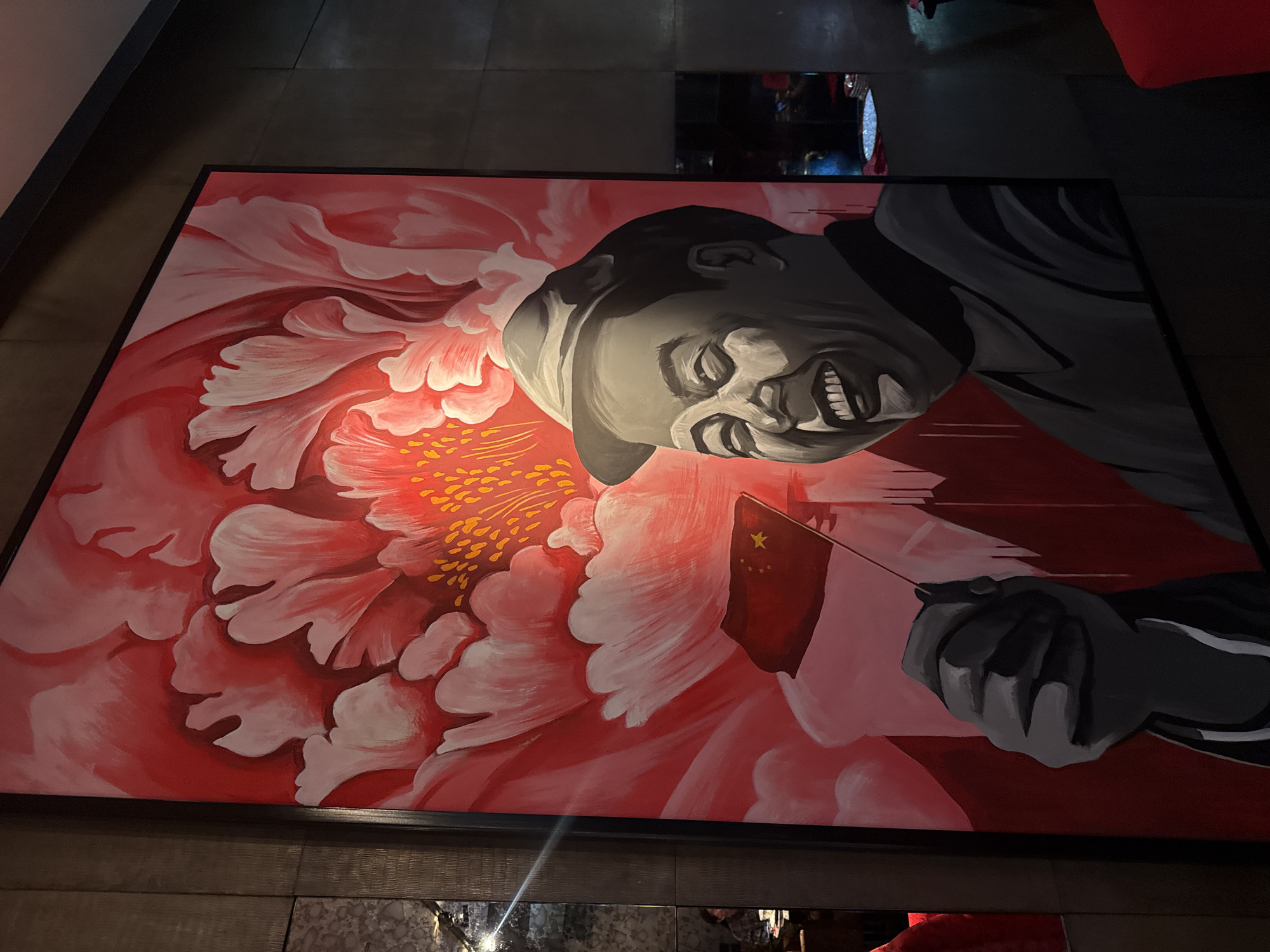 An eye-level, medium shot captures a large, vertically oriented oil painting of a man in a hat, rendered in shades of grey, against a vibrant pink and red floral background. The man, depicted with a wide smile and closed eyes, holds a small Chinese flag in his left hand. The painting is framed in black and hangs on a dark, textured wall. To the left of the painting, a mirrored surface reflects a dimly lit interior with blurred figures and seating arrangements. To the right, the darkness suggests a continuation of the interior space, with glimpses of red seating. The overall lighting is dim, with the painting being the primary source of illumination. The location is identified as Barvikha, Russia.
