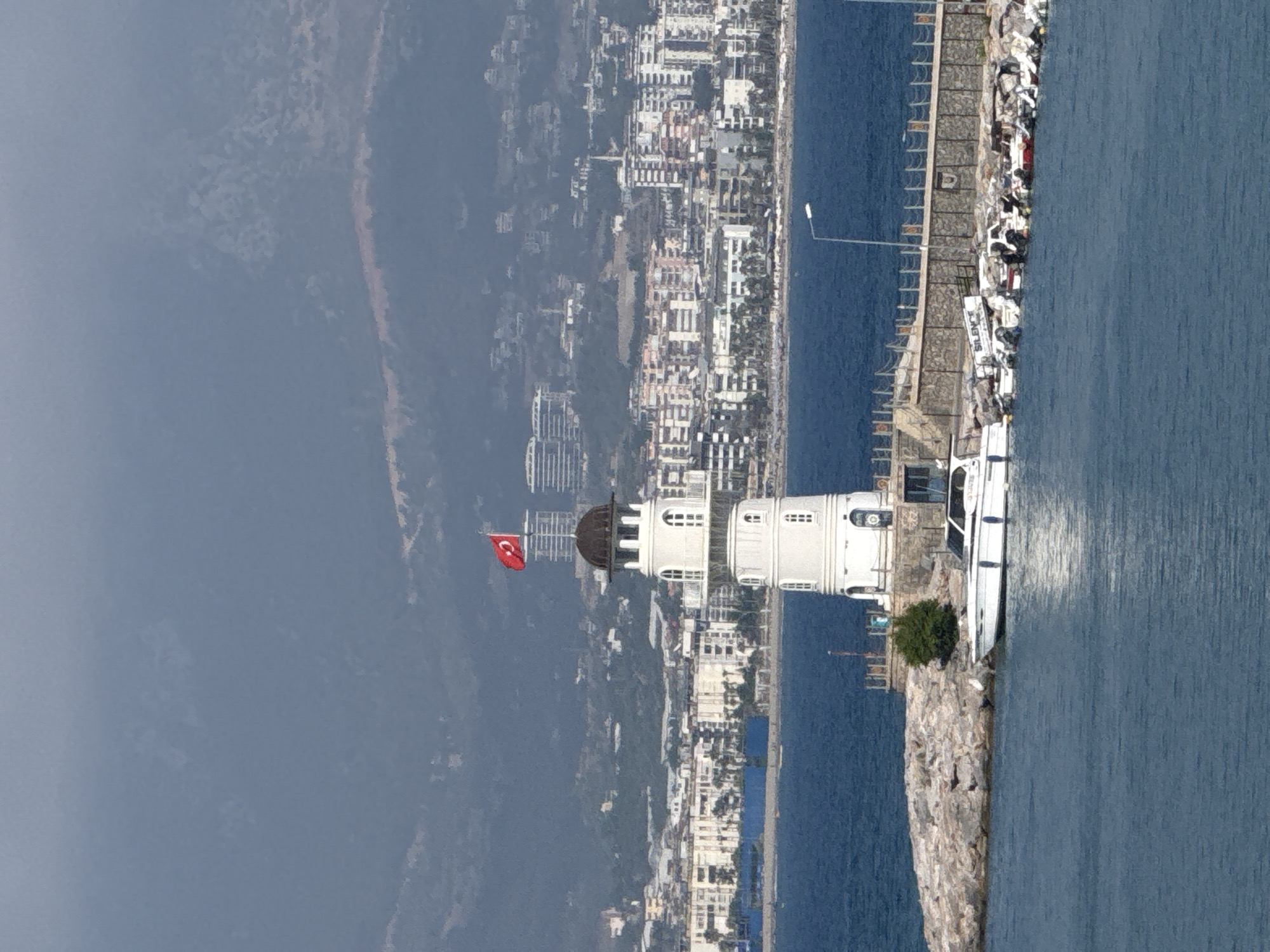 The image shows a white lighthouse on a rocky pier in Alanya, Turkey. The lighthouse is cylindrical with a dark dome and a Turkish flag at the top. The sea is calm and blue, with several boats docked near the lighthouse. In the background, a city skyline with many buildings stretches along the coast, and mountains rise behind the city. The weather appears to be overcast, with a hazy sky.