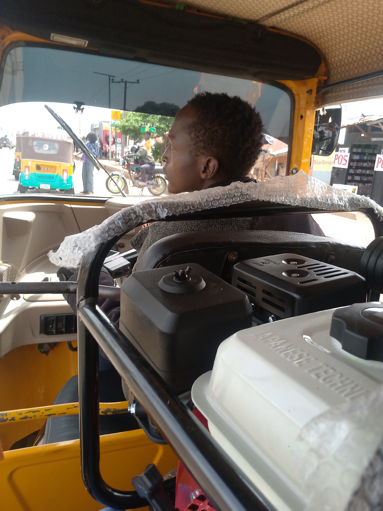 The image depicts the interior of a yellow vehicle, possibly a tricycle rickshaw, in Bukuru, Nigeria. A man with short, dark hair is seated in the driver's seat, looking toward the left. The steering mechanism is encased in bubble wrap. In the foreground is an engine with visible "JAPANESE TECHNIQ" branding and other elements. Through the front windshield, a street scene is visible, including another similar three-wheeled vehicle, pedestrians, and a motorcyclist. A utility pole and commercial signs are also discernible in the background. The vehicle is stationary and in Bukuru, Nigeria.