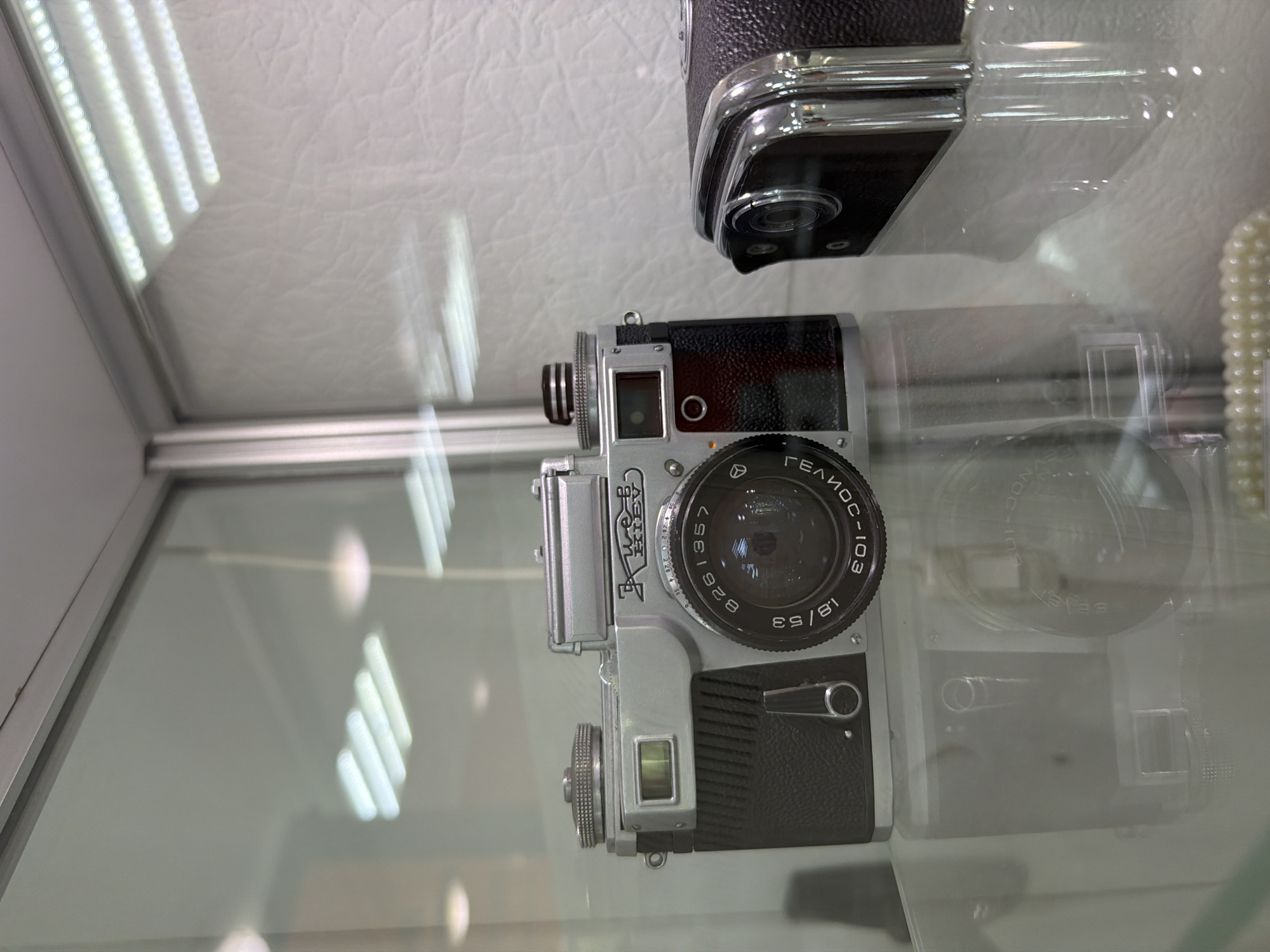 The image depicts a vintage camera, likely a Kiev, displayed in a glass case. The camera is silver and black, with a prominent lens. The background shows a textured white surface and overhead lighting fixtures, suggesting an indoor setting, possibly a museum or a shop. Reflections on the glass indicate the presence of other displays or elements in the environment. The camera's lens displays inscriptions, including "Kiev," "8261357," "1.8/53," and "601-JOC". The overall scene is static and well-lit. The location is identified as Tobolsk, Russia, though no specific landmarks or contextual clues within the image confirm this.
