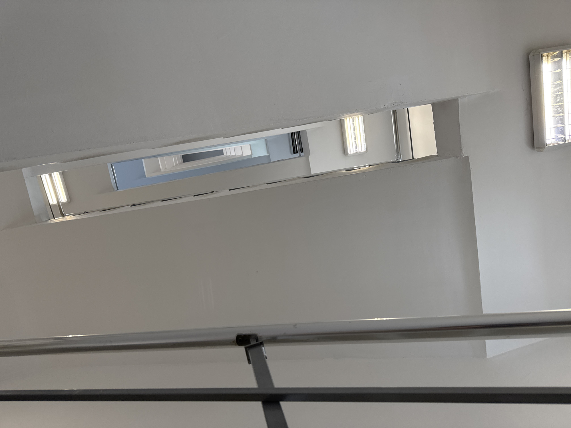 The image depicts the interior of a stairwell in a building. The walls and ceiling are white. The perspective is looking upwards, showing multiple levels of the stairwell receding into the distance. A metal railing is visible in the foreground. There are rectangular fluorescent lights mounted on the walls. The stairwell appears to be located in a city setting, as similar architectural details are present in buildings in Sofia, Bulgaria.