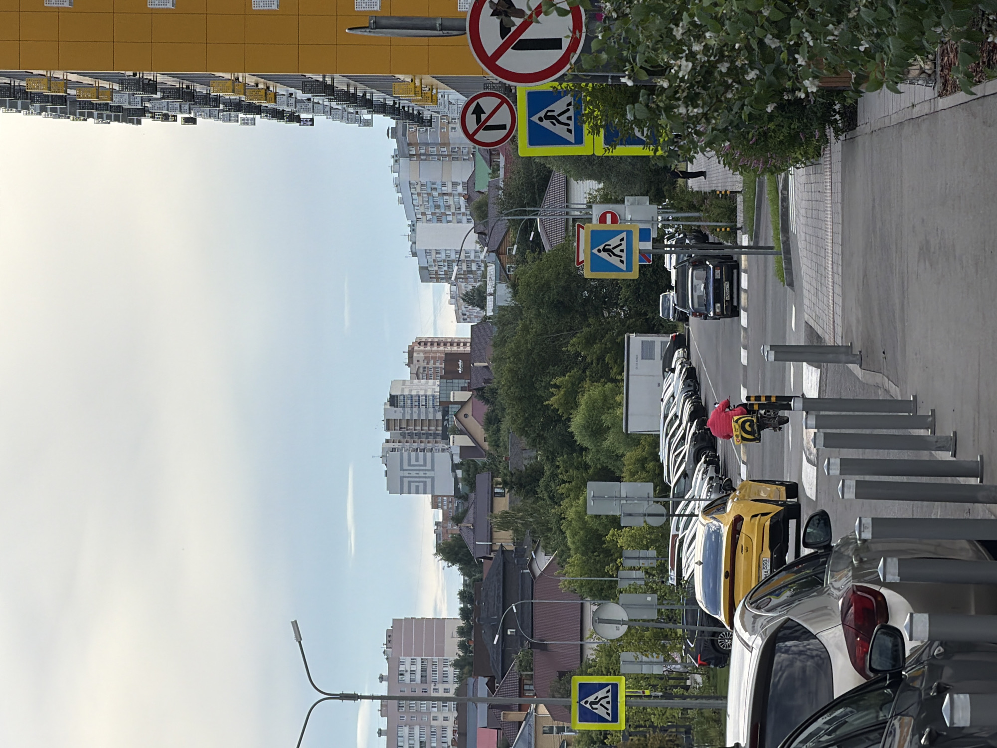A street scene in Kazan, Russia, features a pedestrian crossing with various traffic signs. On the right, a modern yellow building with balconies dominates the foreground. To the left, a row of parked cars, including a yellow Kia, are visible. A person on a scooter with a yellow box on the back is crossing the street. Pedestrian crossing signs and "no turn" signs are present. The sky is overcast.