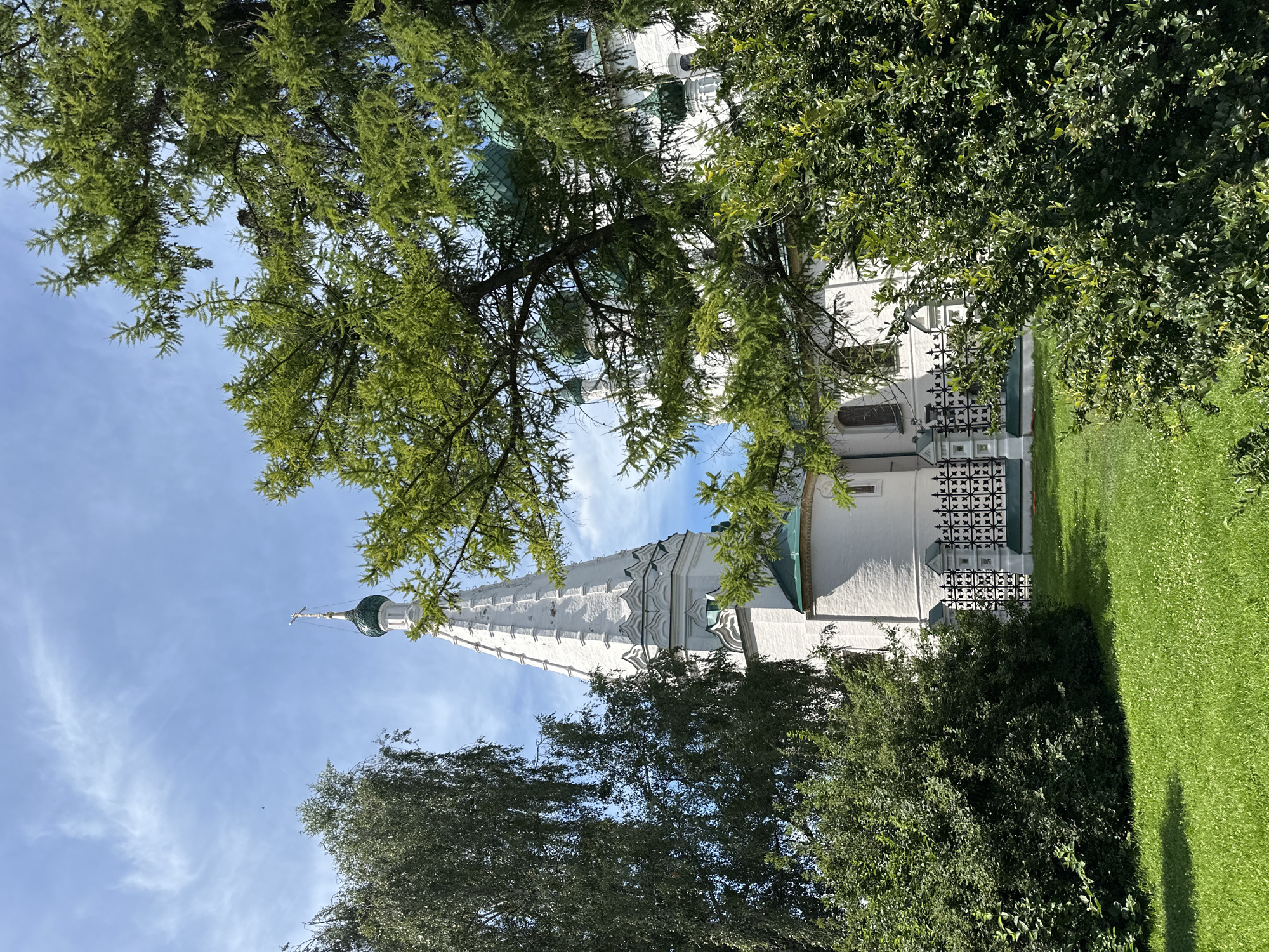 The image depicts an outdoor scene with a white church steeple and a dome visible through lush green trees. The steeple is tall and ornate, with decorative elements and a cross at its apex. The dome is green and rounded. The church itself appears to be constructed of white stone or plaster.
The surrounding area is a well-maintained grassy lawn, with shadows suggesting it is daytime. The sky is a clear blue with some wispy white clouds, indicating pleasant weather. The trees are full of vibrant green leaves, and their branches partially obscure the view of the church, creating a natural framing effect.
There are no people or explicit activities taking place in the image. The focus is on the architectural subject and the natural surroundings. The overall impression is one of tranquility and beauty, likely a serene setting like a church grounds or a historical site. No text is visible in the image. The composition, with the camera angle looking up through the trees towards the steeple, emphasizes the height and grandeur of the church.