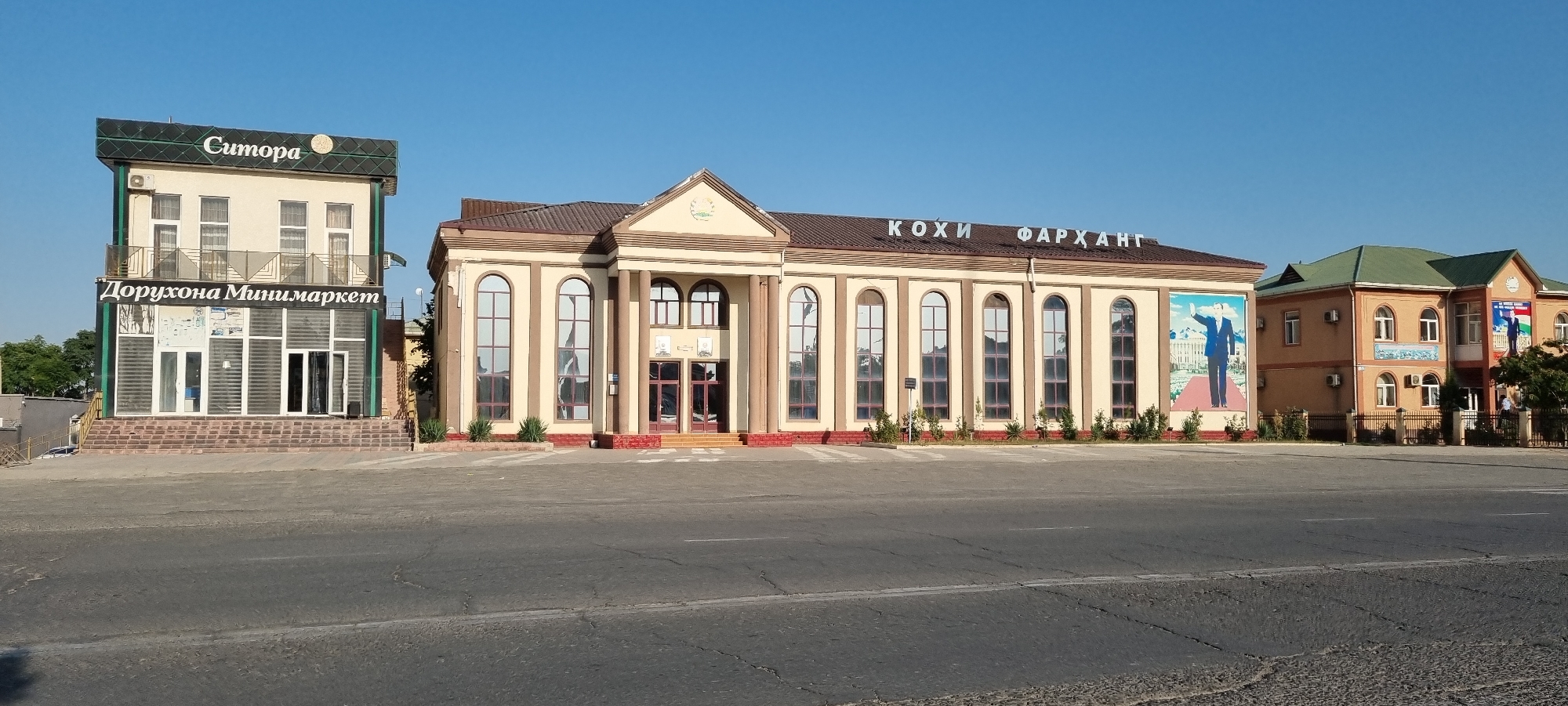 A wide, eye-level shot captures a street scene in Ismoili Somoni, Tajikistan, under a clear, bright blue sky, suggesting a sunny day. The foreground is dominated by a paved road.
In the mid-ground, a row of buildings lines the street. The most prominent building is a large, cream-colored structure with a brown roof, featuring tall, arched windows and classical columns at its entrance. Above the windows, in large, dark letters, the words "КОХИ ФАРХАНГ" are visible. To the left of this building is a narrower, two-story structure, also cream-colored with a dark green trim. The lower floor of this building has large glass windows and doors, with the text "Дорухона Мини-маркет" displayed prominently. The upper floor has several windows and a small balcony. To the right of the "КОХИ ФАРХАНГ" building, another building with a green roof and orange walls is visible, featuring large murals of a man in a suit, possibly a political figure, on its facade.
The overall scene depicts a commercial or public area with businesses and perhaps government buildings. There are no visible people engaged in any activity, nor any strong emotional cues. The clean facades and clear sky suggest a well-maintained environment during daylight hours.