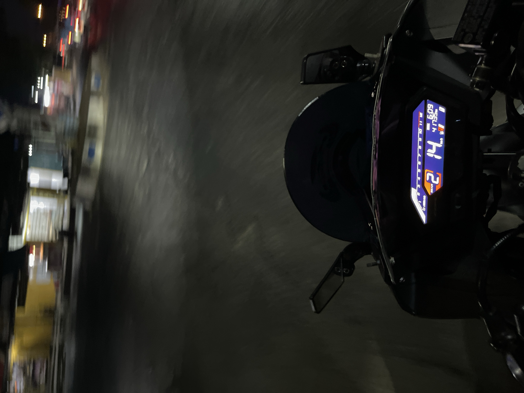 The image captures a first-person perspective of riding a motorcycle at night on a road in Pecangaan, Indonesia. The road surface is dark asphalt, with motion blur indicating movement. In the background, there are blurred lights from buildings and passing vehicles, suggesting an urban or semi-urban environment. Specifically visible are the dashboard of the motorcycle, displaying a digital readout with speed (14 km/h), gear selection (2), time (6:09), and fuel level. The dashboard also shows an odometer reading of 41735. A black windshield is above the dashboard, and a side mirror is visible to the left. The overall scene is characterized by darkness, motion, and the illuminated motorcycle display.