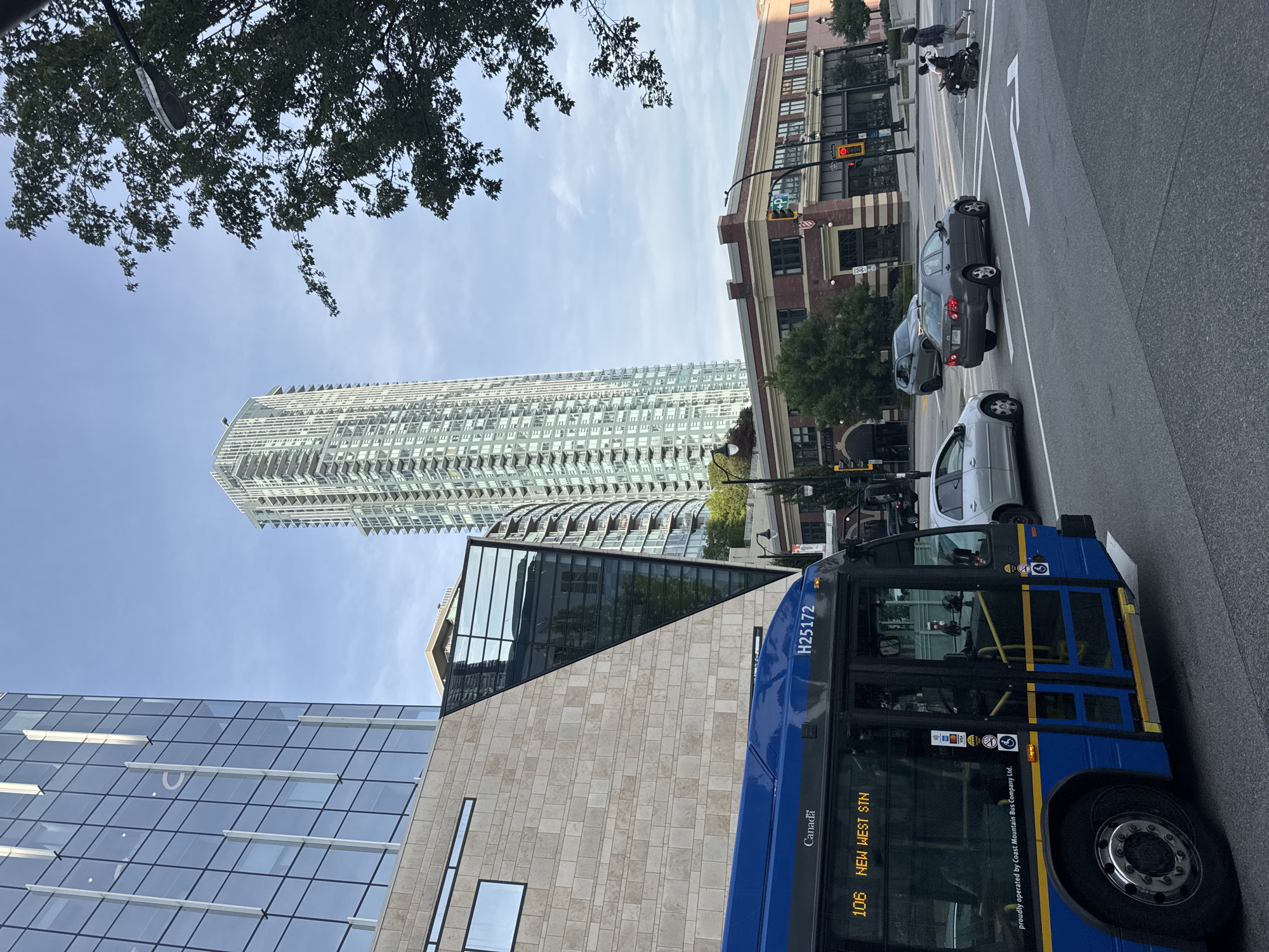 This is a medium shot of a street scene in New Westminster, Canada. The foreground features the side of a blue public transit bus, displaying "106 NEW WEST STN" on its digital sign. The bus is operated by Coast Mountain Bus Company Ltd., as indicated by text on its lower body. To the right of the bus, a gray sedan is stopped at a crosswalk. A person in a wheelchair is being pushed across the street by another person. In the background, a tall, modern skyscraper with numerous balconies dominates the skyline. Adjacent to it are other buildings, including a brick building with arched windows and a glass-fronted building. The sky is partly cloudy and light blue. The scene is set on an urban street with paved roads and visible streetlights.