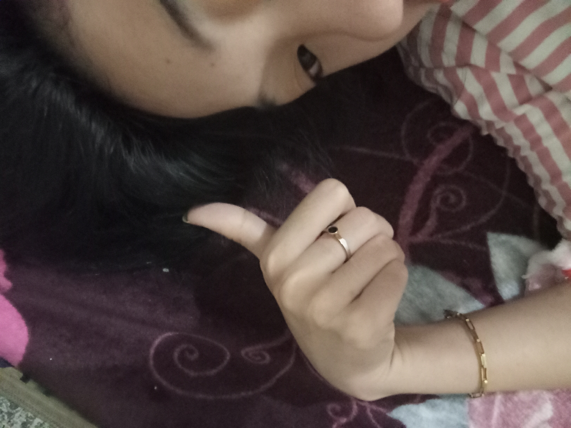 The image shows a close-up of a person's face and hand, resting on a dark purple patterned blanket. The person has black hair and is wearing a ring with a dark stone on their index finger, and a gold bracelet on their wrist. A striped fabric, likely clothing or bedding, is visible in the upper right corner. The scene is indoors, with the lighting suggesting it might be daytime or under artificial light. The specific location in the city of Rejosari, Indonesia cannot be determined from the provided image.