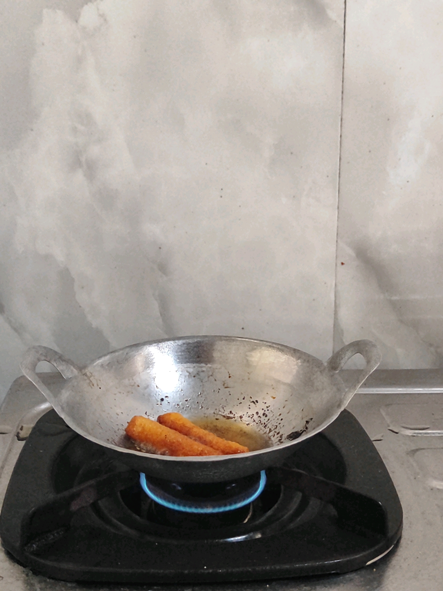 Two fried, breaded items are being cooked in a shallow, metal wok placed on a gas stove. The wok has two loop handles and appears to have some residue from previous cooking. The gas burner beneath the wok is lit, producing a blue flame. The background consists of a light-colored, textured wall. The image is taken from a medium shot, focusing on the cooking process. There are no people present in the scene, nor is there any indication of its specific location within Kotakulon, Indonesia, beyond the general setting of a kitchen.