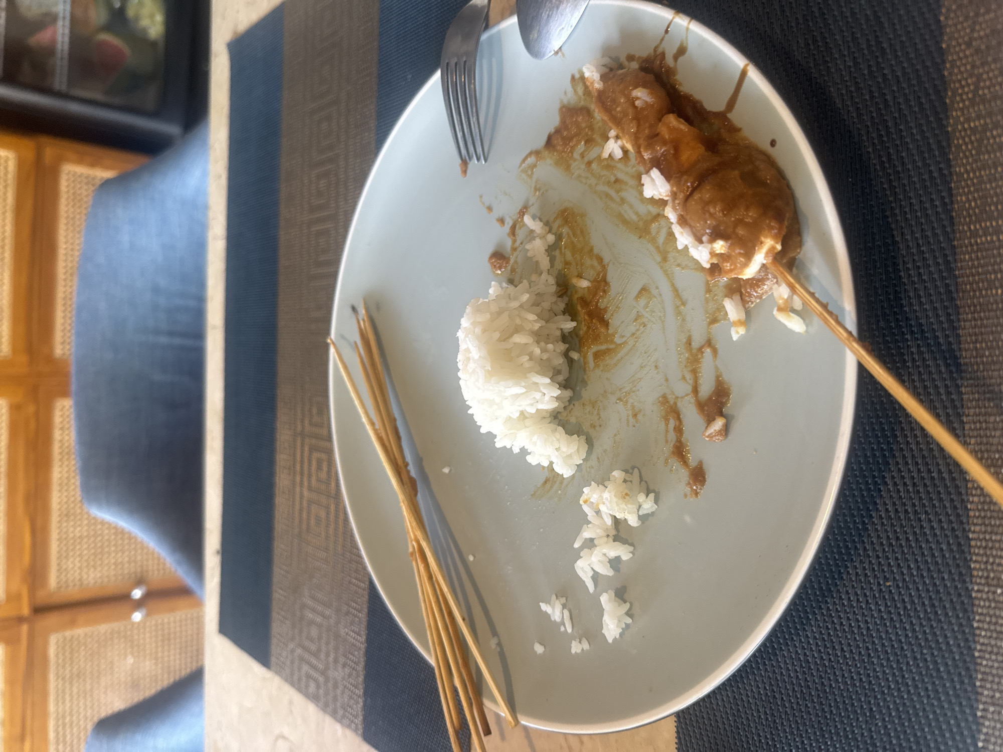 A close-up, overhead shot shows a nearly finished meal on a light blue plate on a table with a woven placemat. The meal consists of rice, satay, and peanut sauce. Several wooden skewers lie to the left of the plate, while one skewer with satay and rice remains on the right. A fork and spoon rest on the right side of the plate. The background shows out-of-focus chairs and wooden cabinetry. This image appears to have been taken in a restaurant in Kuta Selatan, Indonesia.