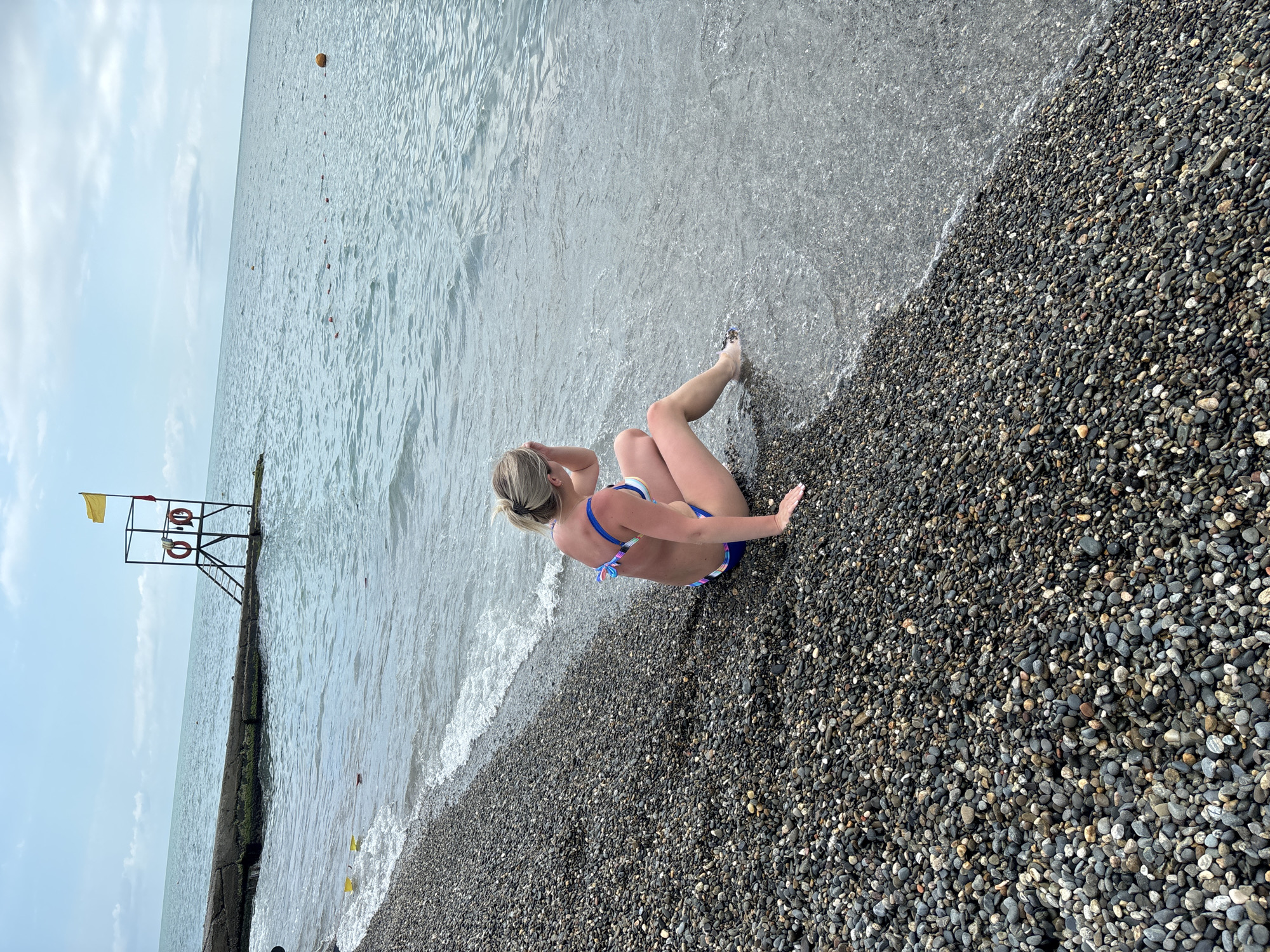 The image shows a woman in a blue and white bikini sitting on a pebble beach with the gentle waves of the sea lapping at her feet. Her back is to the camera, and she is facing the water. She has blonde hair tied up in a bun and her arms are resting on her knees. The beach is covered in dark, smooth pebbles. In the background, a lifeguard tower with a yellow flag and two lifebuoys is visible on a concrete pier. The sky is overcast with white clouds. The overall mood is peaceful and serene. The scene is set on a beach in Sochi, Russia. The time of day appears to be daytime.
