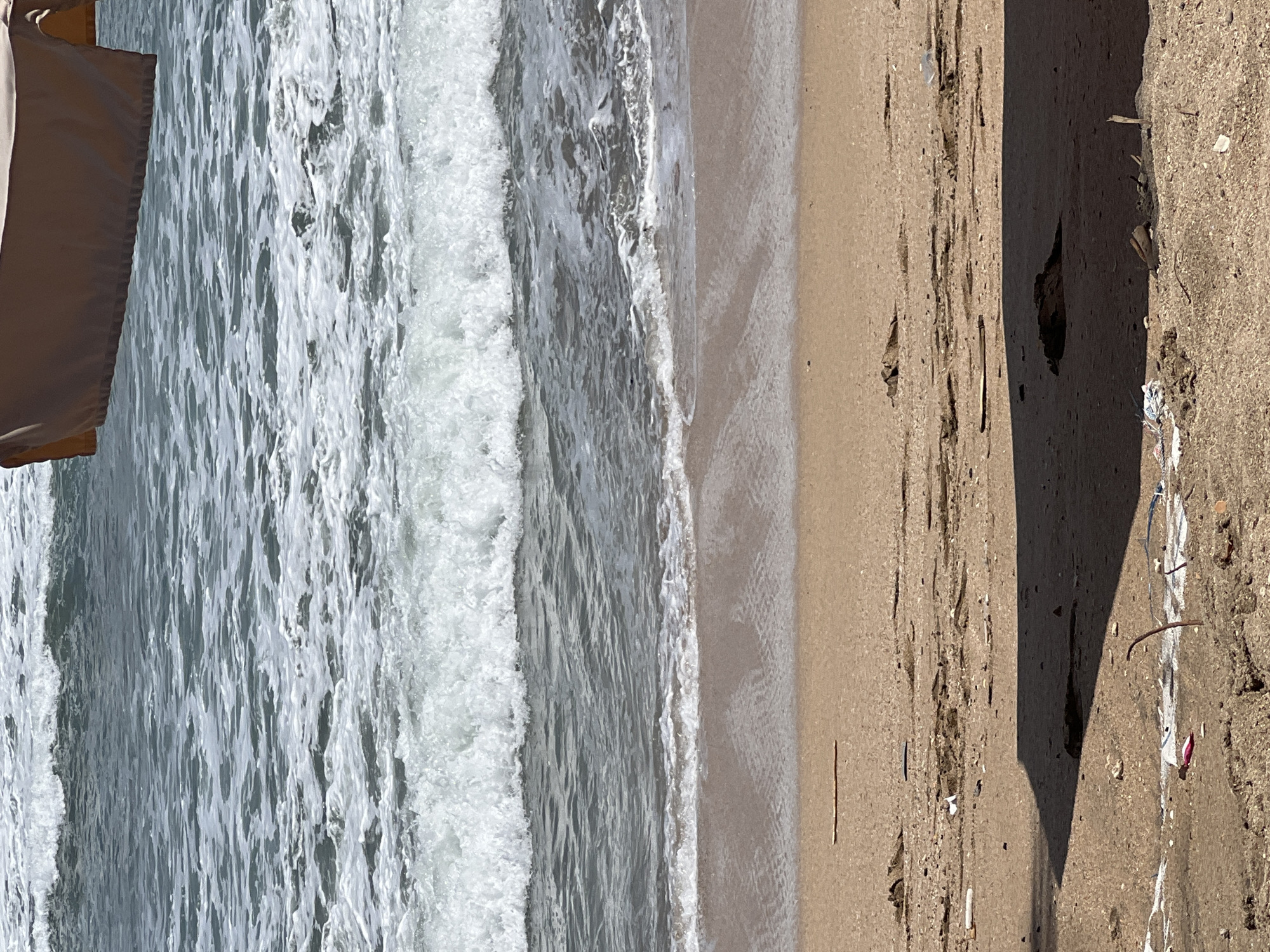 This is an outdoor, eye-level, vertical shot of a beach scene in Kuta, Indonesia. The frame is dominated by the ocean and the shore, with a portion of a light-colored awning in the upper right corner. The ocean is in the upper two-thirds of the image, with waves breaking and forming white foam as they approach the shore. The water is a murky gray-blue. The beach, which occupies the lower third of the image, is a sandy brown color. The sand is littered with debris, including pieces of plastic, driftwood, and other detritus. A dark shadow, cast by an unseen object, falls across the lower portion of the sand. There are no people or animals visible in the scene.