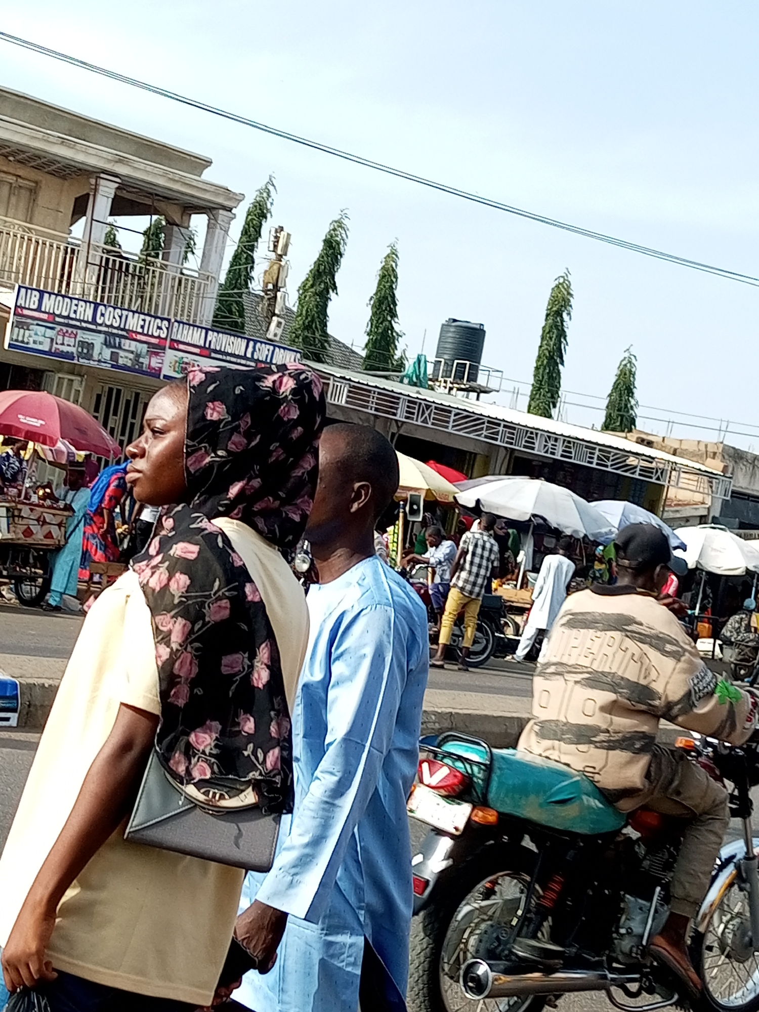 This media file captures a bustling street scene, likely in a market area. The focus is on several individuals and the general environment.
In the foreground, a woman wearing a light yellow garment and a black and pink floral headscarf is seen from the side. She is carrying a light grey clutch bag. Beside her, a man in a light blue outfit is also partially visible. Further back, a person is riding a motorcycle, wearing a tan and black patterned jacket.
The background reveals a street lined with shops and stalls, indicated by awnings and various goods displayed. There are also several tall, slender trees lining the street. A large black water tank is visible on top of one of the buildings. The sky is bright and clear, suggesting it is daytime, likely mid-morning or afternoon, with pleasant weather.
Visible text on a building reads "AIB MODERN COSTMETICS RAHAMA PROVISION & SOFT DRINKS". The overall impression is one of everyday life and commerce in a busy urban setting. The individuals appear to be going about their daily activities, with no strong emotions evident. The scene suggests a vibrant local market or commercial district.