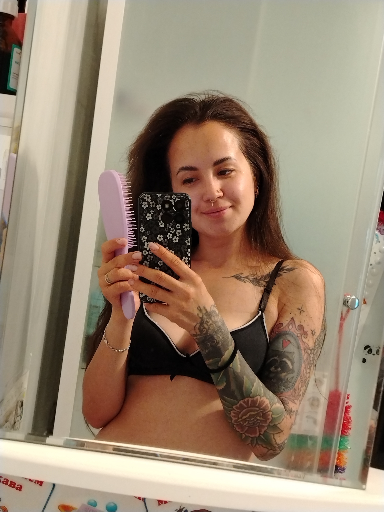A woman with long, dark hair and tattoos on her arms is taking a mirror selfie in a bathroom. She is wearing a black bra with white trim and holding a purple hairbrush in her left hand and a smartphone in her right hand. The smartphone has a black case with a floral pattern. The background includes a mirror, a shower curtain, and some toiletries on a shelf. The image appears to have been taken in a residential bathroom in Noviy, Russia.