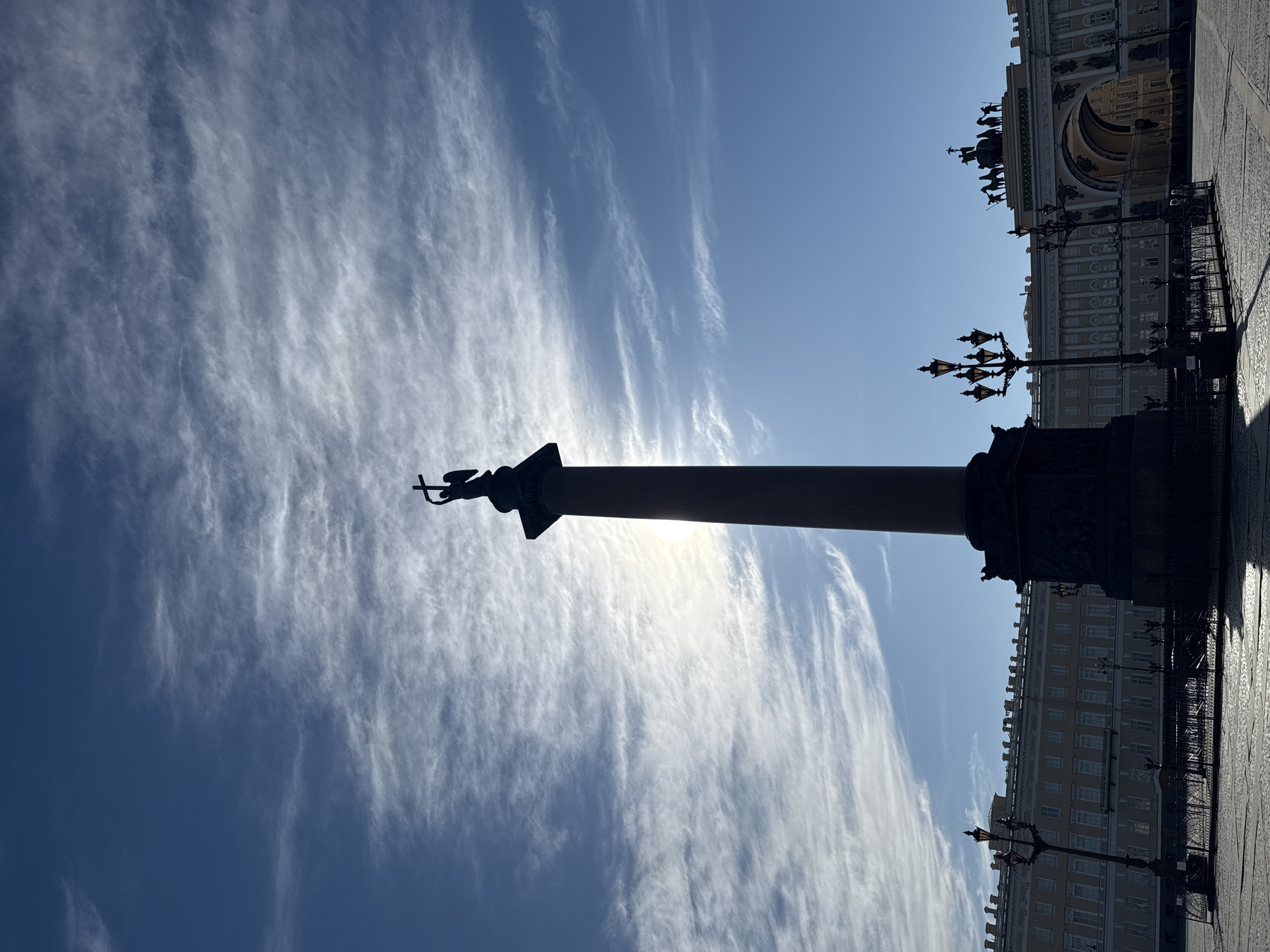 The image captures a monumental outdoor scene in Saint Petersburg, Russia, featuring the iconic Alexander Column and a portion of the General Staff Building in Palace Square. The primary subject is the Alexander Column, which stands tall and is heavily silhouetted against a bright, sunlit sky. At its apex, the sculpture of an angel holding a cross is discernible.
To the right of the column, the grand architecture of the General Staff Building is visible, also largely in silhouette. Its distinctive triumphal arch is clearly identifiable, topped with a sculpture of horse-drawn chariots. Several ornate street lamps, dark against the bright background, are positioned in the mid-ground. The ground below appears to be paved with cobblestones or large flagstones, reflecting some light in patterns.
The sky is a vibrant blue, adorned with dramatic, wispy cirrus clouds that stretch across the frame, indicating movement in the upper atmosphere. The sun is positioned behind the Alexander Column and the buildings, creating a strong backlighting effect that renders most of the foreground and architectural elements as deep, dark silhouettes. This intense sunlight suggests either early morning or late afternoon, with the sun relatively low in the sky, though bright enough to illuminate the clouds. There is no discernible human activity or visible text in the image. The overall impression is one of grandeur, historical significance, and serene monumentality under a dynamic sky.