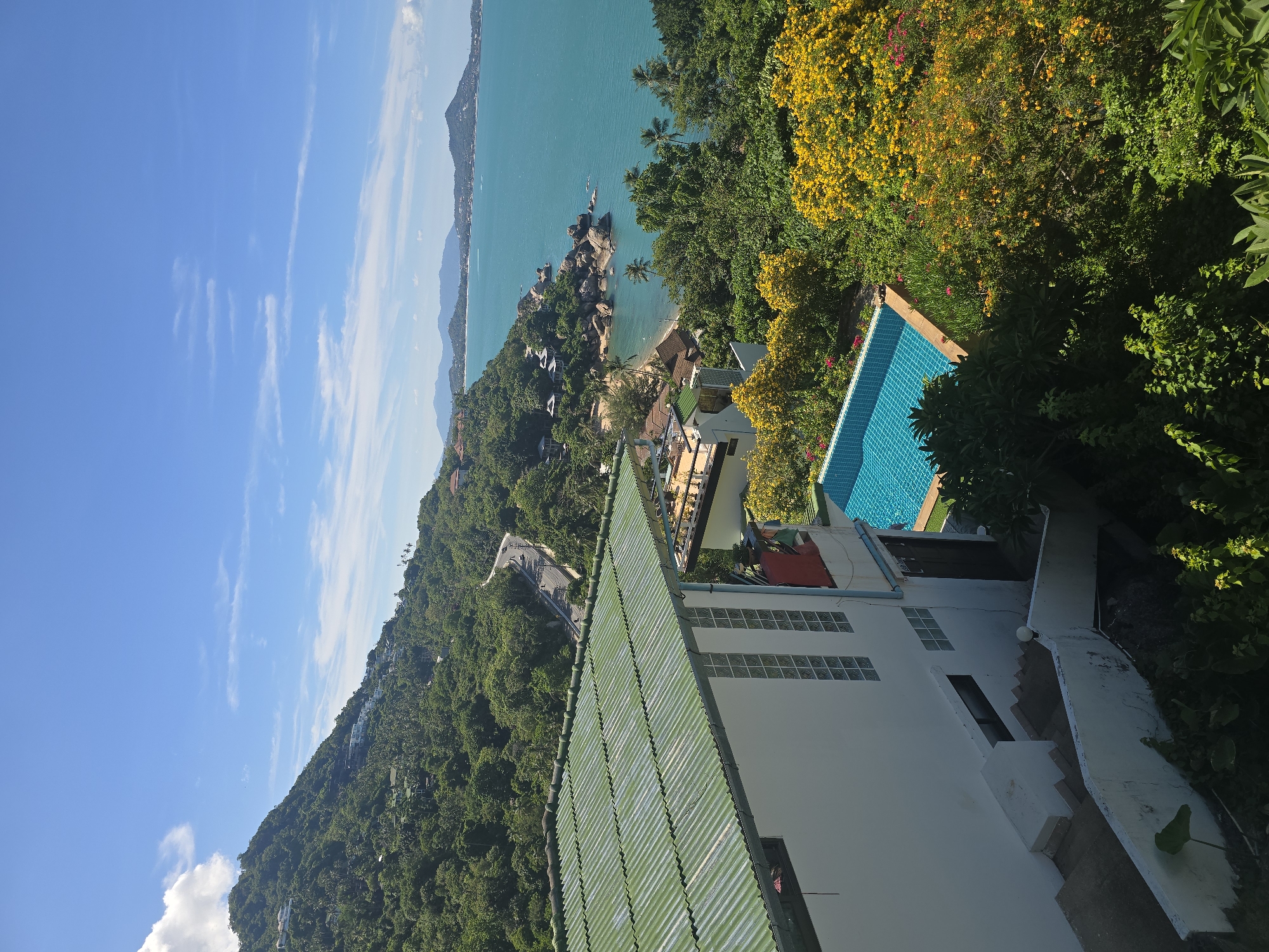 This is a photograph of a scenic coastal landscape taken from an elevated viewpoint. The foreground features part of a building with a light-colored exterior and a green corrugated metal roof. Adjacent to this building is a bright blue swimming pool, partially visible. Lush green trees and vibrant yellow flowers with some pink blooms are scattered throughout the scene, creating a colorful contrast with the blue of the ocean.
In the mid-ground, the landscape slopes down towards a turquoise ocean. A small, sandy beach is nestled amongst large, grey rocks along the coastline. Several small buildings or structures are visible amongst the trees on the hillside overlooking the water.
The background shows a hazy coastline with mountains in the distance. The ocean stretches out to the horizon under a clear blue sky with scattered white, wispy clouds. The lighting suggests it is daytime, likely mid-morning or afternoon, with bright sunlight casting shadows. The overall impression is one of a tropical or Mediterranean location, offering a peaceful and idyllic view. No people or specific activities are clearly depicted.