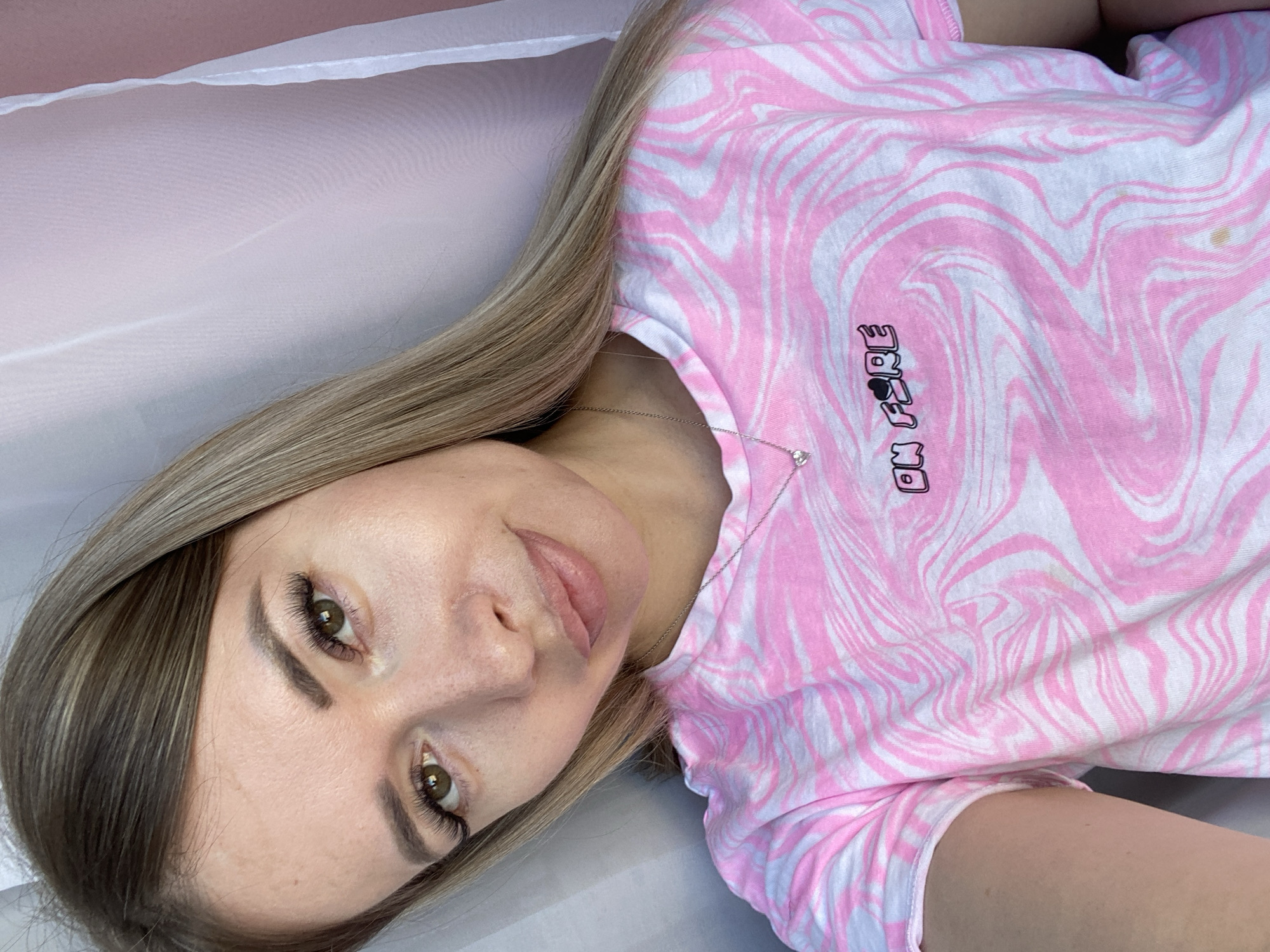 A woman lies on her back on a white, possibly fabric-covered surface. She wears a pink and white patterned t-shirt with the phrase "ON FIRE" written on it. She is fair-skinned with blonde hair, defined eyebrows, long eyelashes, and a silver necklace with a small pendant. The image is angled, with the woman's face towards the lower-left corner. The city location is not identifiable from the provided image.