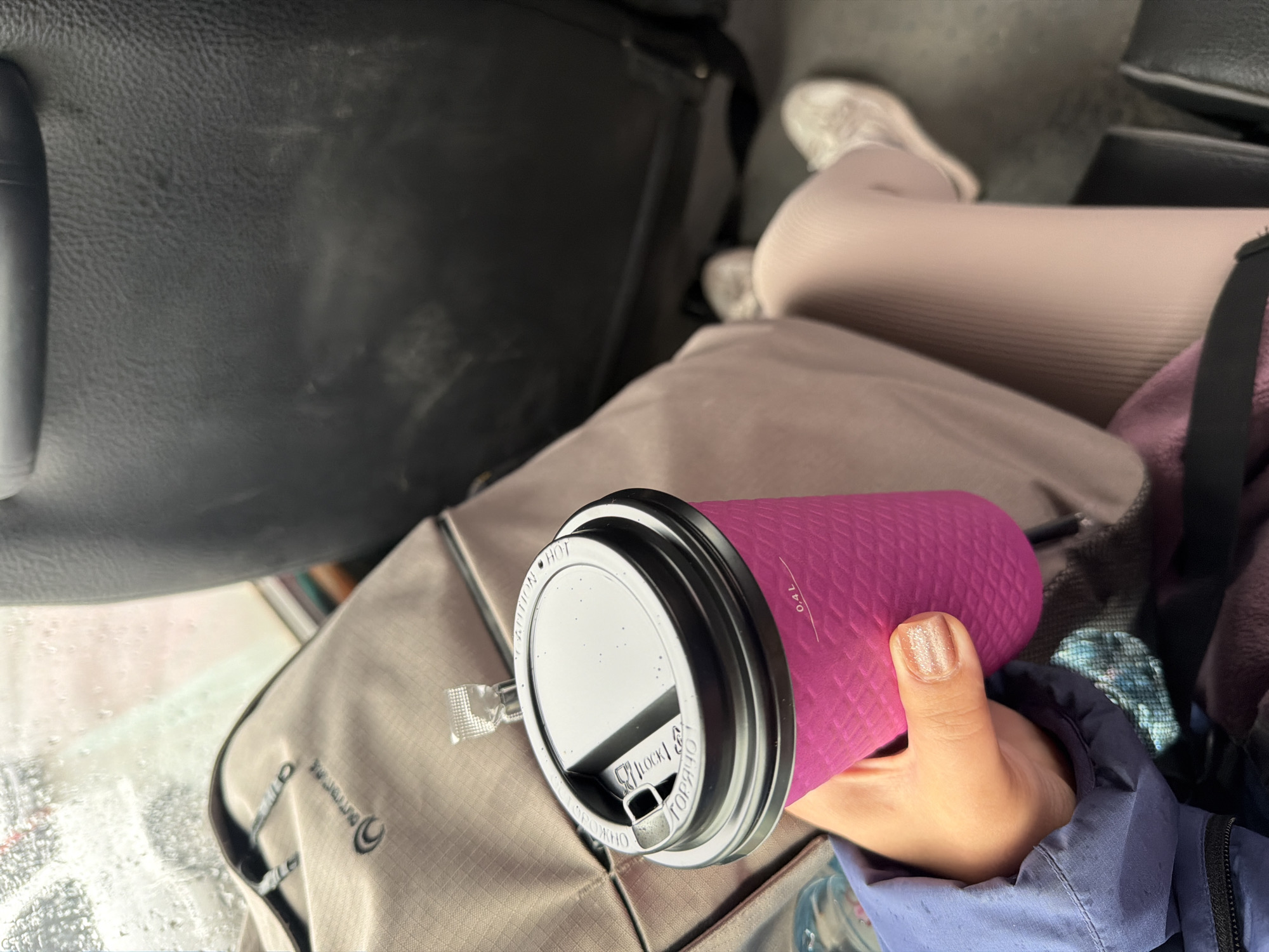 The image depicts a person holding a pink, textured disposable coffee cup with a black lid. The cup has a visible measurement of "0.4 L" on its side. The person's hand, with glitter nail polish, is gripping the cup. They are wearing a blue jacket. In the background, a light brown backpack with a logo is visible, along with a dark seat. The scene appears to be inside a vehicle, possibly a bus or train, as indicated by the window with water droplets and the seating arrangement. The location is specified as Petropavlovsk-Kamchatsky, Russia.