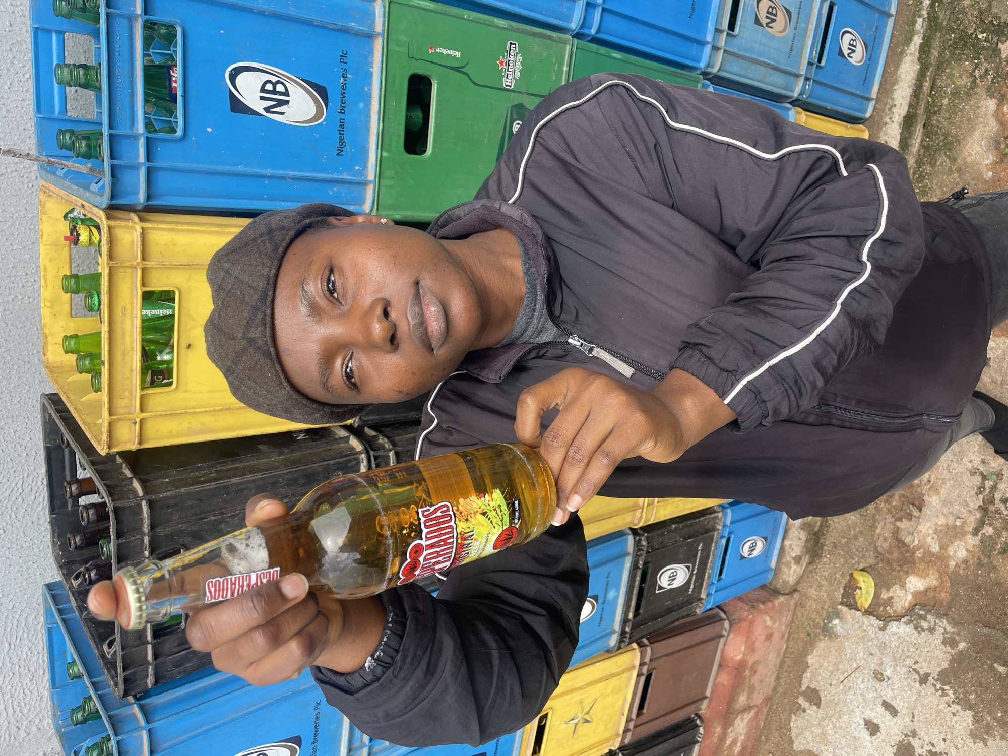 This image captures a person holding a bottle of beer in an outdoor setting, likely a beverage storage area or retail point in Jos, Nigeria.
The central subject is a person with dark skin, wearing a dark cap and a black track jacket with white piping on the sleeves. They are looking directly at the camera with a neutral expression, their gaze steady and composed. They hold a clear glass bottle of "DESPERADOS ORIGINAL BEER," identifiable by its prominent red "DESPERADOS" lettering, presenting it towards the viewer with both hands. The bottle contains a golden-amber liquid with a visible foam head near the bottleneck.
The background consists of numerous colorful plastic crates stacked, suggesting a commercial or industrial context related to beverages. The crates are primarily blue, yellow, and green, with some darker ones. Many of these crates contain empty green glass bottles. Visible text on the crates includes "NB," "Nigerian Breweries Plc," and "Heineken," indicating the presence of products from these brands. The ground appears to be a worn concrete surface, and the natural lighting suggests the photo was taken during the day. The scene depicts a casual moment within a functional, everyday environment associated with drink distribution or sales.