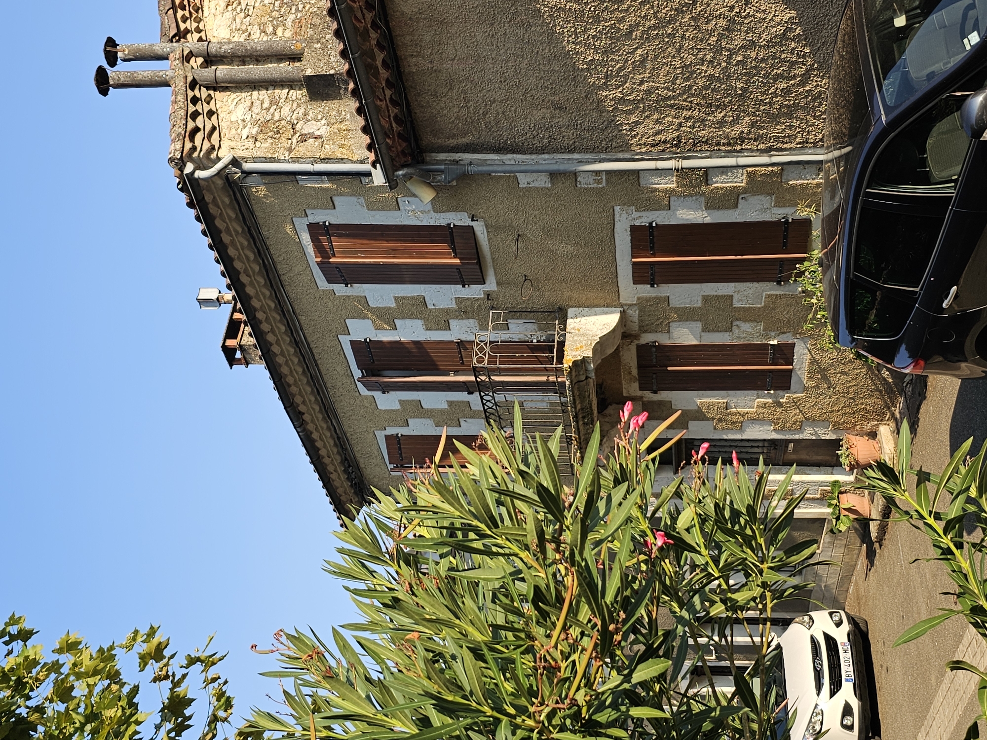 This is a medium shot, eye-level, outdoor photograph taken during the day in Lavardens, France. The image features a traditional two-story building with a stucco exterior painted a light yellow, accented with white stone detailing around the windows and corners. The building has a terracotta tiled roof and two prominent chimney stacks. Several windows are visible, all fitted with dark brown wooden shutters. A small wrought-iron balcony is present on the second floor, above a doorway.
In the foreground and to the left, a large, leafy green bush with small pink flowers partially obscures the view of the house. In front of the house, a white SUV is parked, its license plate reading "BY - 402 - MD". To the right of the SUV, two potted plants are visible on the ground. A portion of a dark-colored car is visible on the far right of the frame, at the bottom. The sky is clear and blue.