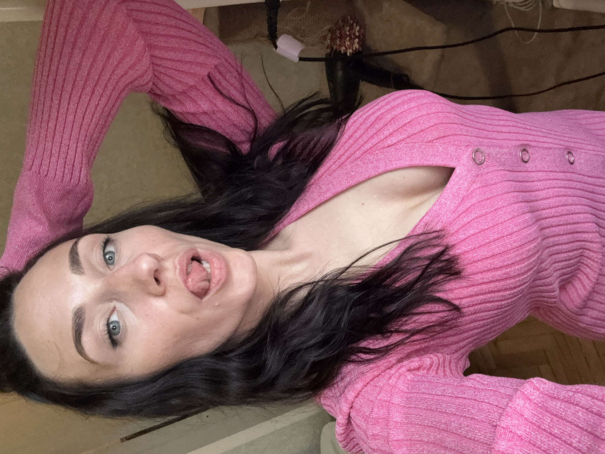 A woman with fair skin, dark hair, and light blue eyes lies on what appears to be a floor. She is wearing a pink ribbed sweater with a low neckline. Her mouth is open, and her tongue is slightly protruding. The background includes a cluttered area with a hairbrush and electrical cords. There is no indication of the location being Saint Petersburg, Russia, or any specific city.