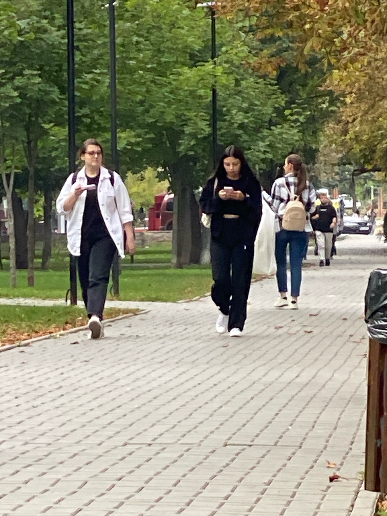 The image depicts a pedestrian walkway paved with gray rectangular bricks. Several individuals are walking along this path. In the foreground, a person with glasses wearing a white open shirt over a black t-shirt and black pants is walking towards the viewer, holding a white object in their left hand. To their right and further down the path, a young woman with long dark hair, dressed in a black long-sleeved top and black pants, is looking down at her phone. Behind her, another young woman with her hair tied back, wearing a plaid shirt and jeans, carries a light-colored backpack. Further in the background, other individuals and vehicles are visible, suggesting an urban environment. Trees with green and autumn-colored foliage line the walkway. The lighting indicates it is daytime. The overall scene is consistent with a street or park pathway in Chișinău, Moldova, given the context provided.