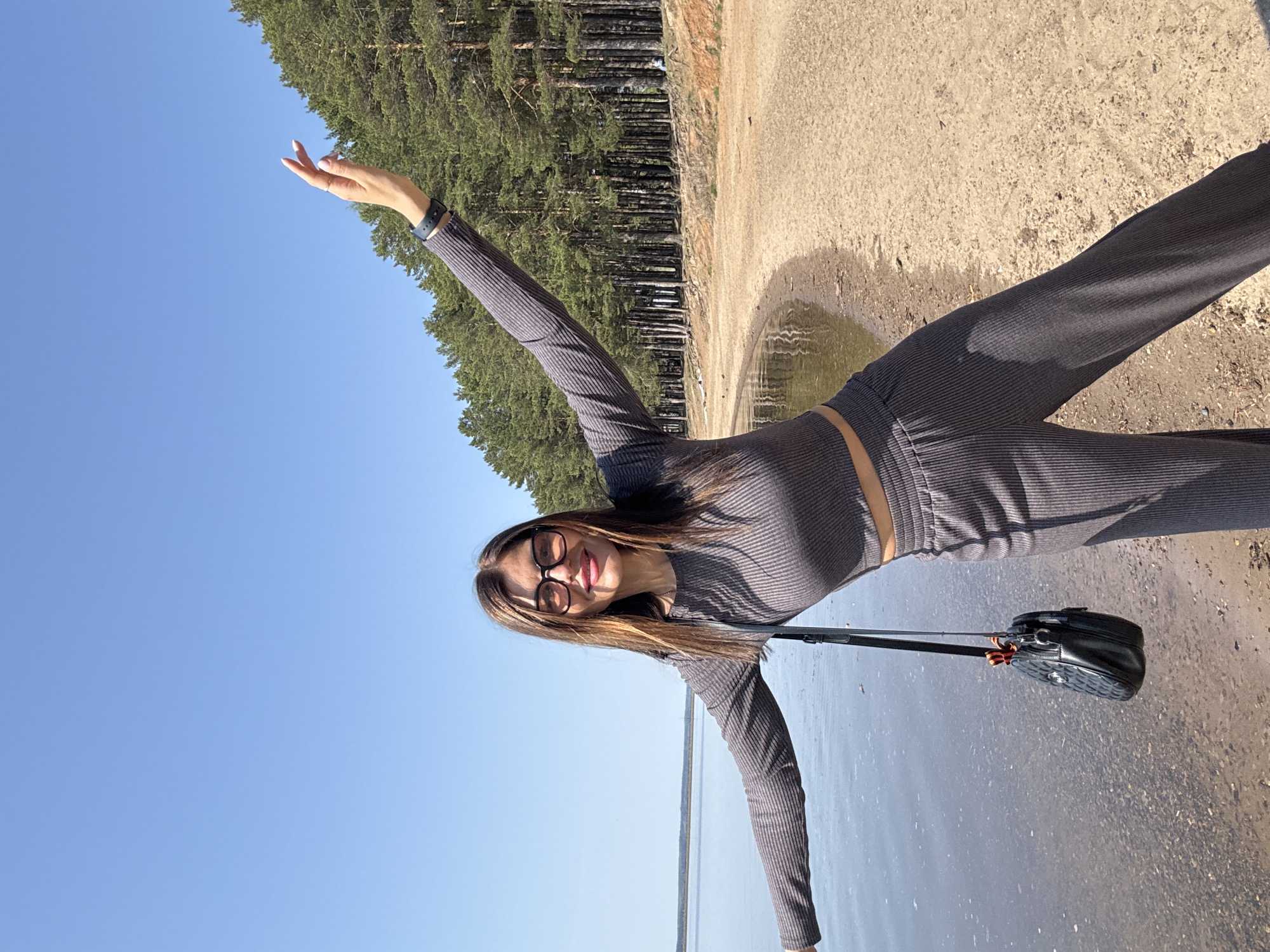 A woman stands on the beach in Chaikovsky, Russia. She is wearing glasses and a gray, ribbed two-piece outfit, with one arm raised towards the sky and a purse over her shoulder. Behind her, a lake extends to the horizon under a clear blue sky. A forest of tall pine trees lines the edge of the beach to the left. The ground is sand and a small puddle of water.