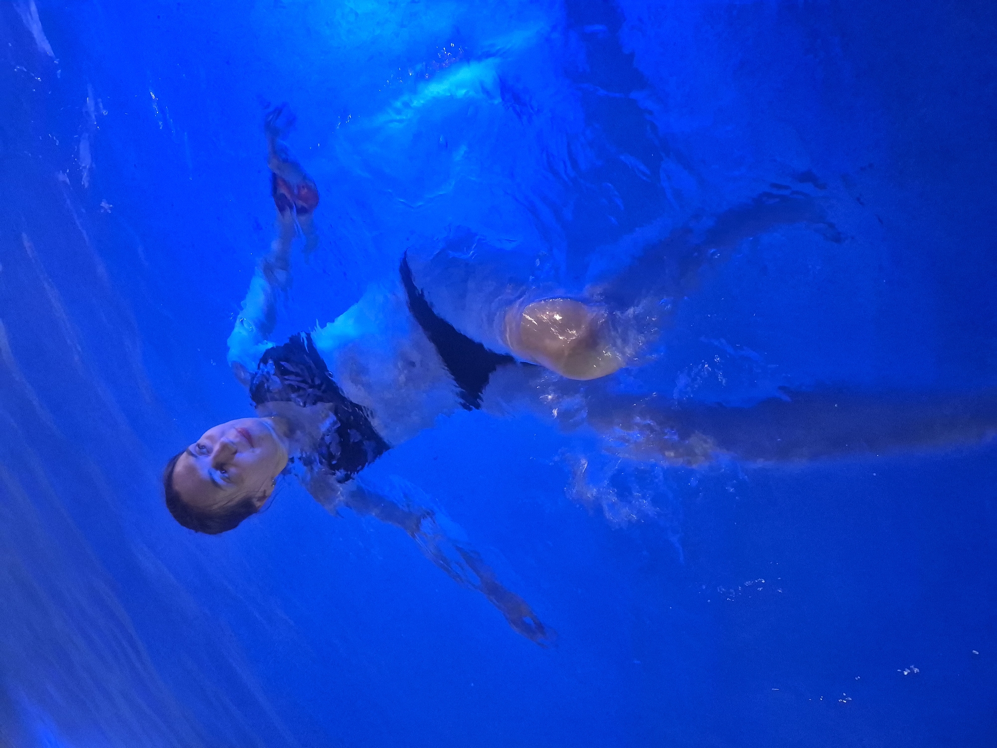 An overhead shot depicts a person, likely a woman, floating on her back in a vibrantly blue body of water, suggestive of a swimming pool. Her head is tilted back, with her face looking upwards, revealing a calm and serene expression. She is wearing a dark-colored swimsuit, visible on her upper torso and hips. Her arms are slightly outstretched to her sides, and her legs are extended beneath the water, creating subtle ripples and air bubbles around her.
The water is intensely illuminated with a consistent blue light, giving it a deep, almost luminous quality. This strong blue hue dominates the entire frame, with lighter reflections and patterns visible on the surface and around the submerged parts of the body. The perspective offers a top-down view of the person gracefully suspended in the water. The activity portrays a moment of relaxation and weightlessness.
The scene conveys a tranquil atmosphere, emphasized by the person's peaceful expression and the uniform blue environment. The location context indicates this event is taking place in Votkinsk, Russia. There is no visible text present in the image.