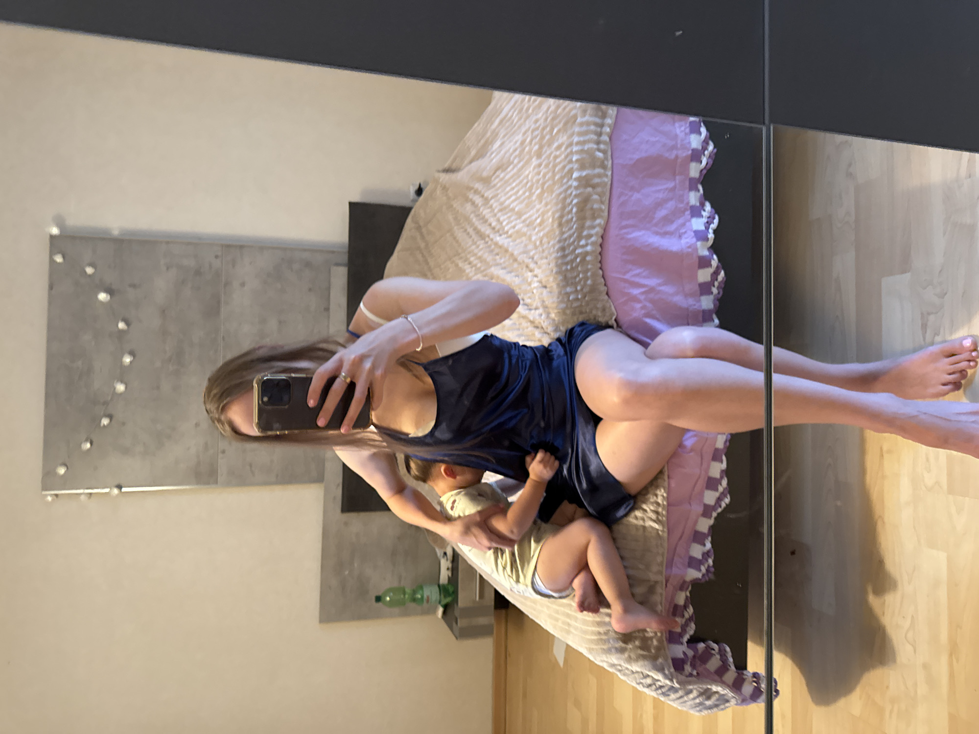 In Überherrn, Germany, a woman is captured in a mirror selfie taken in a bedroom. She is sitting on the edge of a bed, dressed in a dark blue satin slip dress and a white bra. Her right hand is holding her phone, and her left arm is around a young child who is leaning against her. The child is wearing a light-colored t-shirt and appears to be lying down, facing away from the camera. The bed is covered with a beige textured blanket and a lilac-colored bed skirt. To the left of the bed, on a nightstand, sits a green bottle of water. The room features a light-colored wall and a large mirror reflecting the scene. The floor is made of light-colored wood.