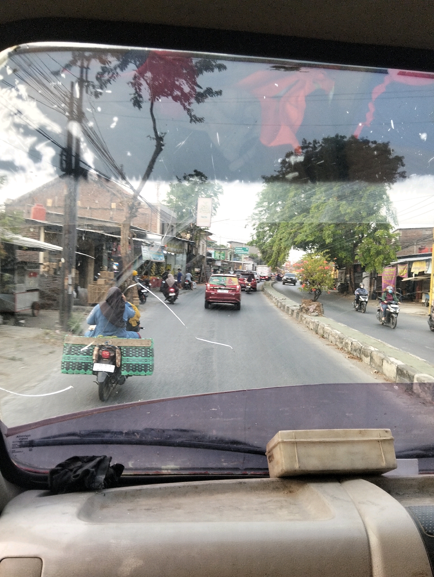 This is a medium-distance, eye-level, outward-facing, medium shot of a street scene in Semarang, Indonesia, taken from inside a vehicle. The perspective is slightly angled and blurry, as if seen through a dirty windshield. In the foreground, a worn, beige plastic container sits on top of the dashboard. A black cloth, possibly gloves, is draped over the left side of the dashboard.
The middle ground features a road with traffic. A motorcyclist wearing a blue garment and a headscarf is riding a motorcycle with a large, green, caged cargo carrier attached to the back. Several other motorcycles and cars are visible on the road, some moving and some parked. Buildings line the side of the street, some appearing to be shops or houses. Trees and utility poles are also present.
The background shows more buildings and trees, and the sky is overcast. The overall impression is of a typical day on a busy street in a Southeast Asian city. The image is characterized by its lack of sharp focus, the presence of smudges and scratches on the "windshield" (likely the lens of the camera), and a slightly desaturated color palette.