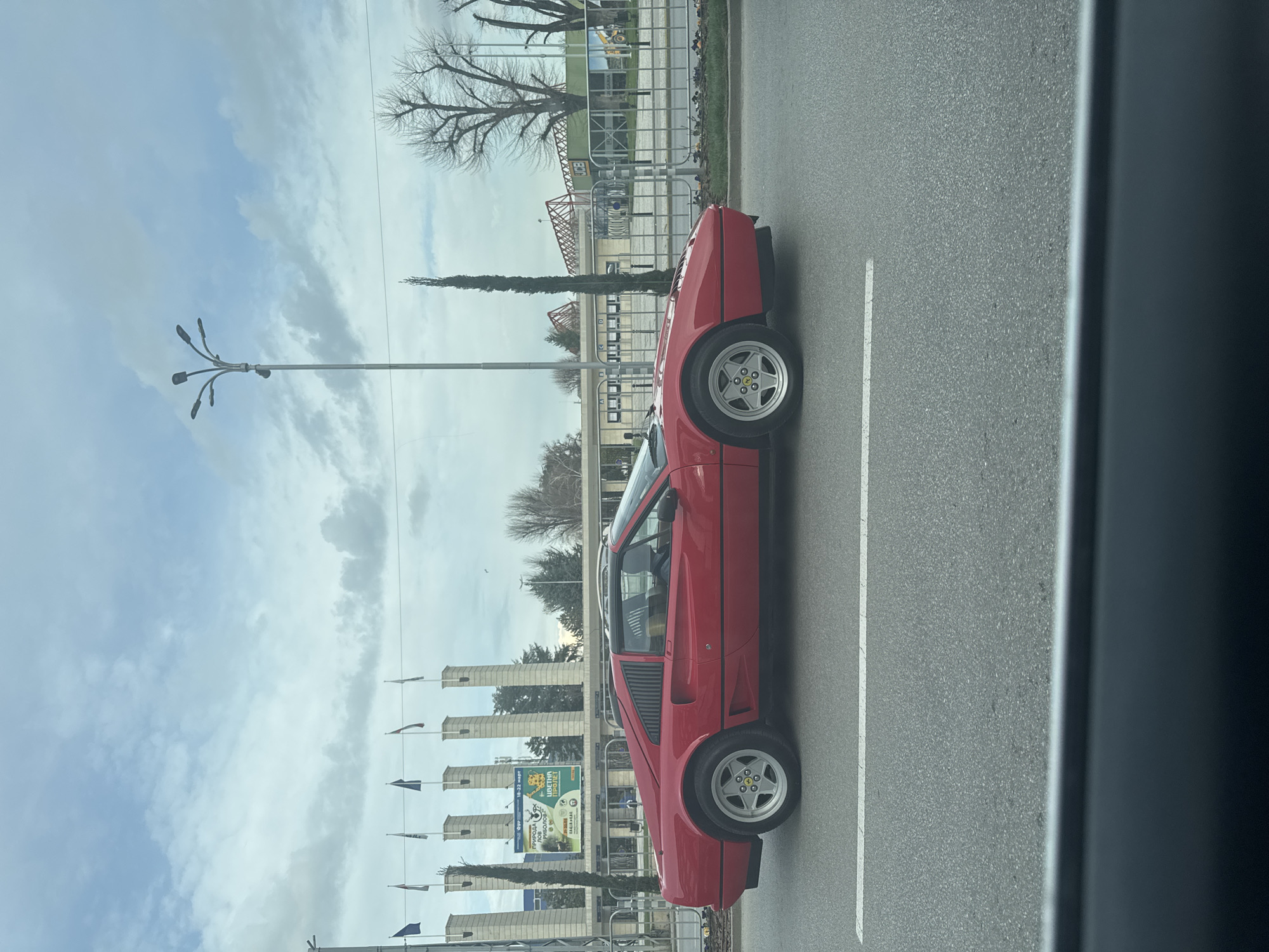 A red Ferrari car sits on a road next to the International Fair Plovdiv in Plovdiv, Bulgaria. The car faces the right. The road has a white lane divider. In the background, the fairground's entrance pillars with flags, trees, and other structures are visible under a cloudy sky. The image is taken from inside a vehicle, with a dark grey interior visible on the right.