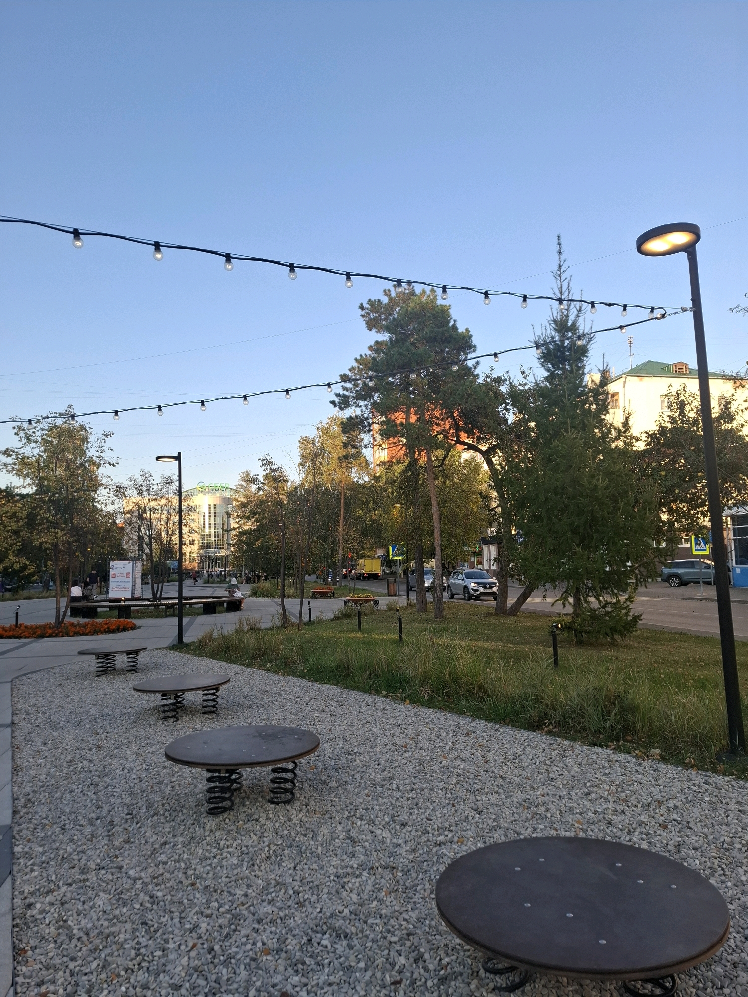 The image depicts an outdoor public space in Tyumen, Russia. In the foreground, there are several round tables with spring bases, scattered across a gravel pathway. Beyond the pathway, there is a grassy area with trees and decorative orange flowers. A street lamp with an illuminated head stands prominently on the right side of the image, and string lights with bulbs are stretched across the sky above. In the background, buildings, cars on a street, and more trees are visible under a clear blue sky. The scene appears to be a park or a public square.