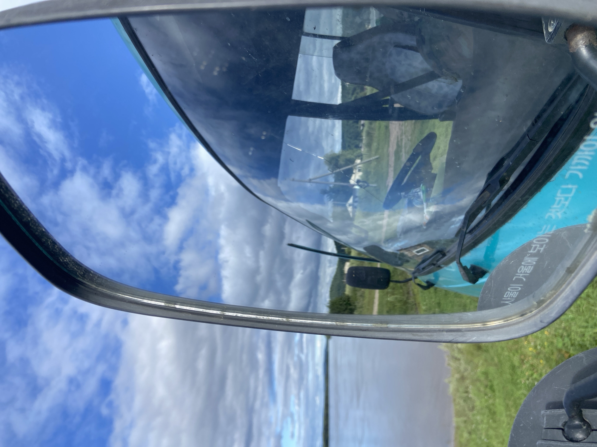 The image shows a view from inside a vehicle, looking into a side mirror. The mirror reflects a cloudy blue sky and a body of water. The vehicle's windshield is also visible, with a view of the interior, including a steering wheel and a seat. The exterior of the vehicle appears to be light blue. The environment suggests a rural or natural setting, possibly near a river or lake. There are no people visible in the image, nor any specific indications of the city of Черняево, Russia. The scene captures a moment of travel, with the focus on the reflections and the surrounding landscape.