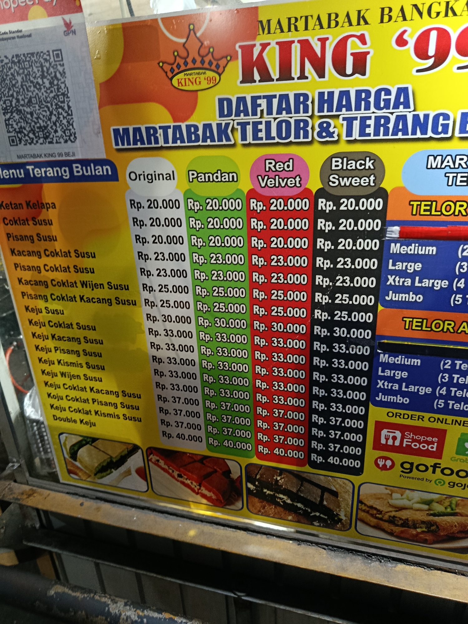 The image is a close-up, eye-level shot of a brightly colored menu board for "MARTABAK KING '99" in Depok, Indonesia. The menu advertises "MARTABAK BANGKA" and lists prices for "MARTABAK TELOR & TERANG BULAN" (fried pancake and sweet martabak) in various flavors including Original, Pandan, Red Velvet, and Black Sweet. The prices range from Rp. 20.000 to Rp. 40.000. There are also sections for "TELOR" and "TELOR A" with sizes like Medium, Large, Xtra Large, and Jumbo, accompanied by parenthetical notes that are partially obscured but appear to indicate quantities of fillings or ingredients.
Below the pricing sections, there are images of martabak. One image shows a slice of martabak with a dark filling, possibly chocolate, layered with cream. Another image depicts a close-up of a martabak cut into squares, revealing a thick, creamy filling with green elements, possibly durian or pandan.
The lower right portion of the board displays logos for "Shopee Food" and "gofood", indicating that orders can be placed online through these platforms. The text "Powered by gojo" is also visible. A QR code is present on the left side of the board, labeled "MARTABAK KING 99 BEJI". The overall impression is of a vibrant, appealing advertisement for a popular Indonesian street food. The lighting suggests it is daytime, possibly in an outdoor or semi-outdoor setting.