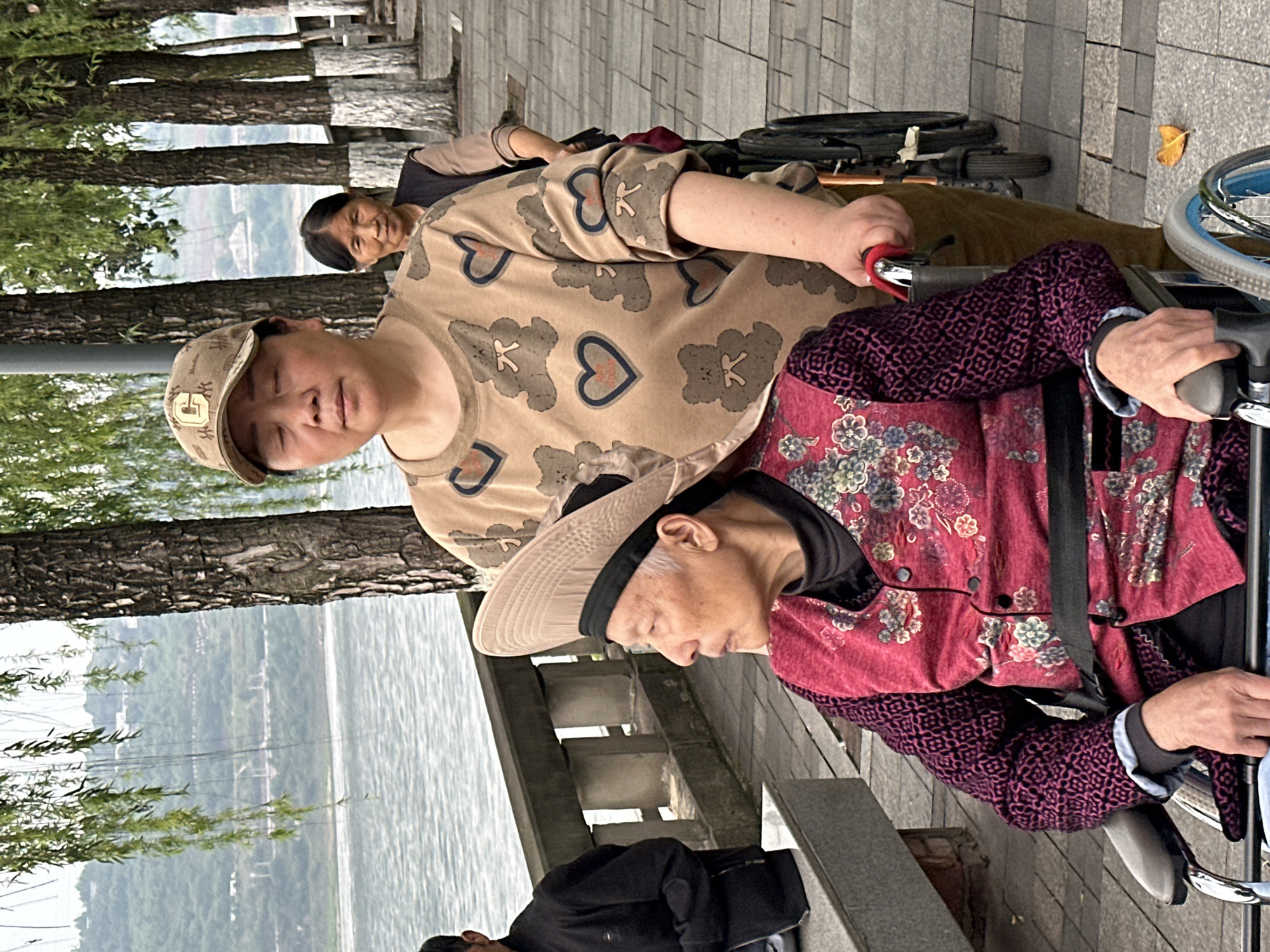 The photograph depicts a woman pushing a wheelchair along a paved path, with an elderly woman seated in the wheelchair. Another woman follows behind them. The location appears to be Jiangjin, China, as suggested by the landscape and the style of dress of the people in the photograph. The path is adjacent to a body of water, possibly a river or lake, and lined with trees.