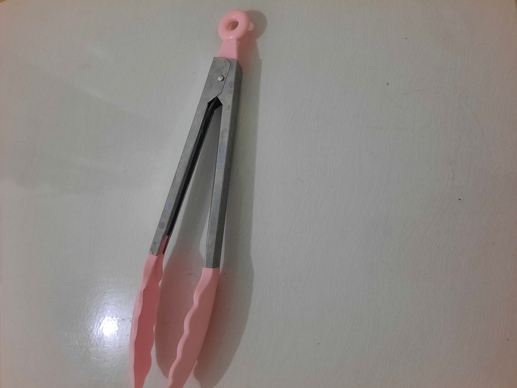 A pair of pink silicone tipped tongs is lying open on a light-colored, textured surface. The tongs are made of stainless steel and have a loop at the end for hanging. The surface appears to be a tabletop or counter, possibly in a kitchen. The lighting in the image suggests an indoor setting, with a bright light source reflecting off the surface. There are no people or other objects visible in the frame. There is no information in the image to indicate the location in Lhokseumawe, Indonesia.