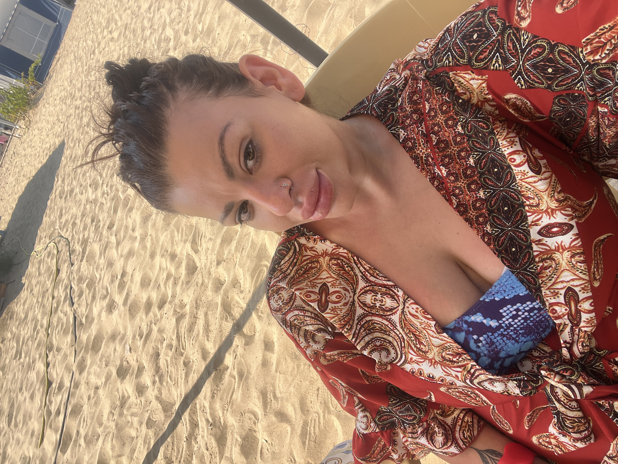 A woman is lying on a chair on a sandy beach in Sozopol, Bulgaria. She is wearing a red and patterned robe over a blue, snake-print bikini top. The sand is light brown and shows footprints and some debris. In the background, a blue and white tent structure and some green foliage are visible. The sun is casting a shadow across the sand from an unseen object.