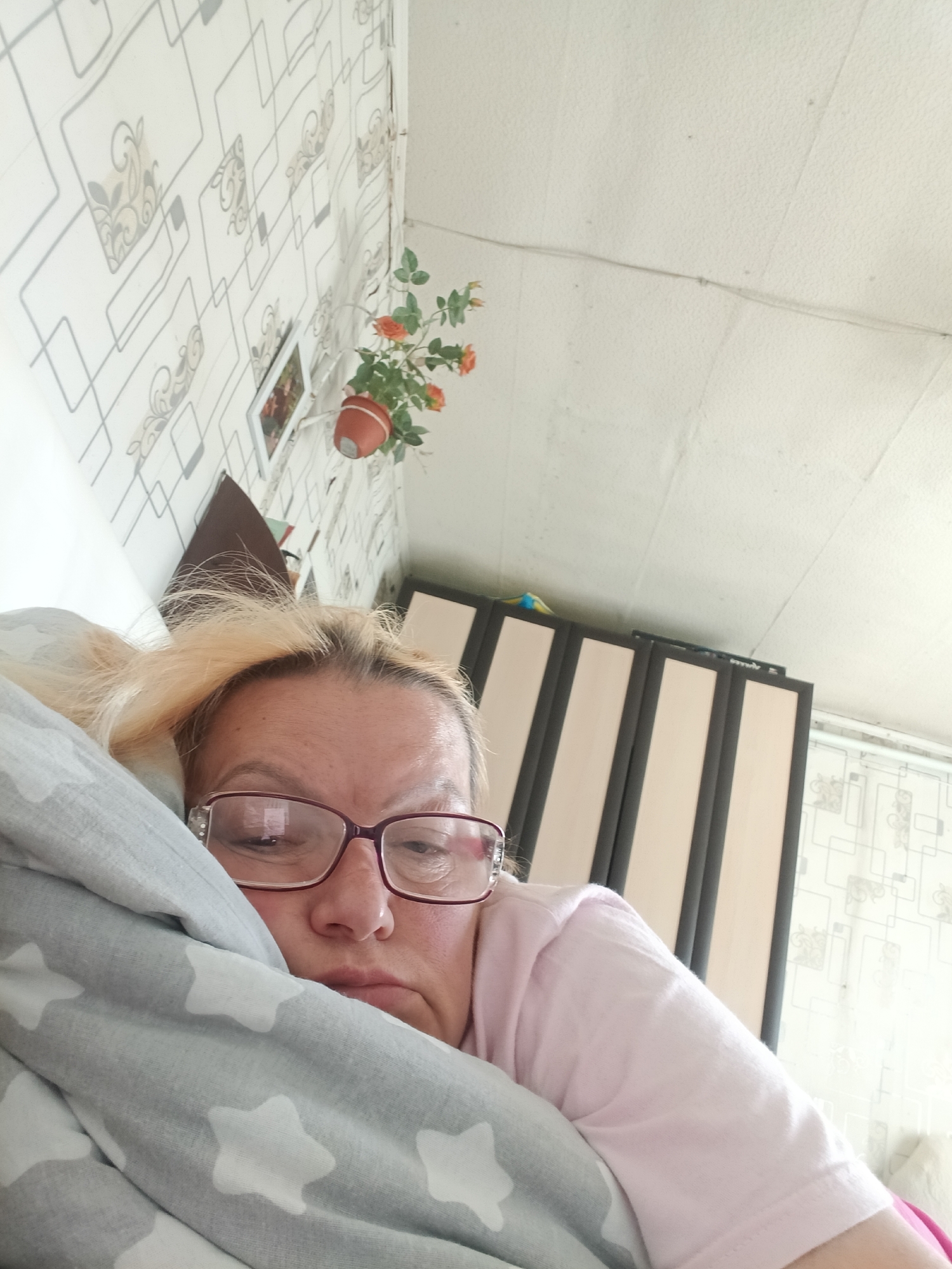 A woman with blonde hair and glasses lies in bed, partially covered by a grey pillow with white stars. She wears a light pink shirt. The background includes patterned wallpaper, a hanging plant in a terracotta pot, and a wardrobe with vertical stripes. The location is a domestic interior.