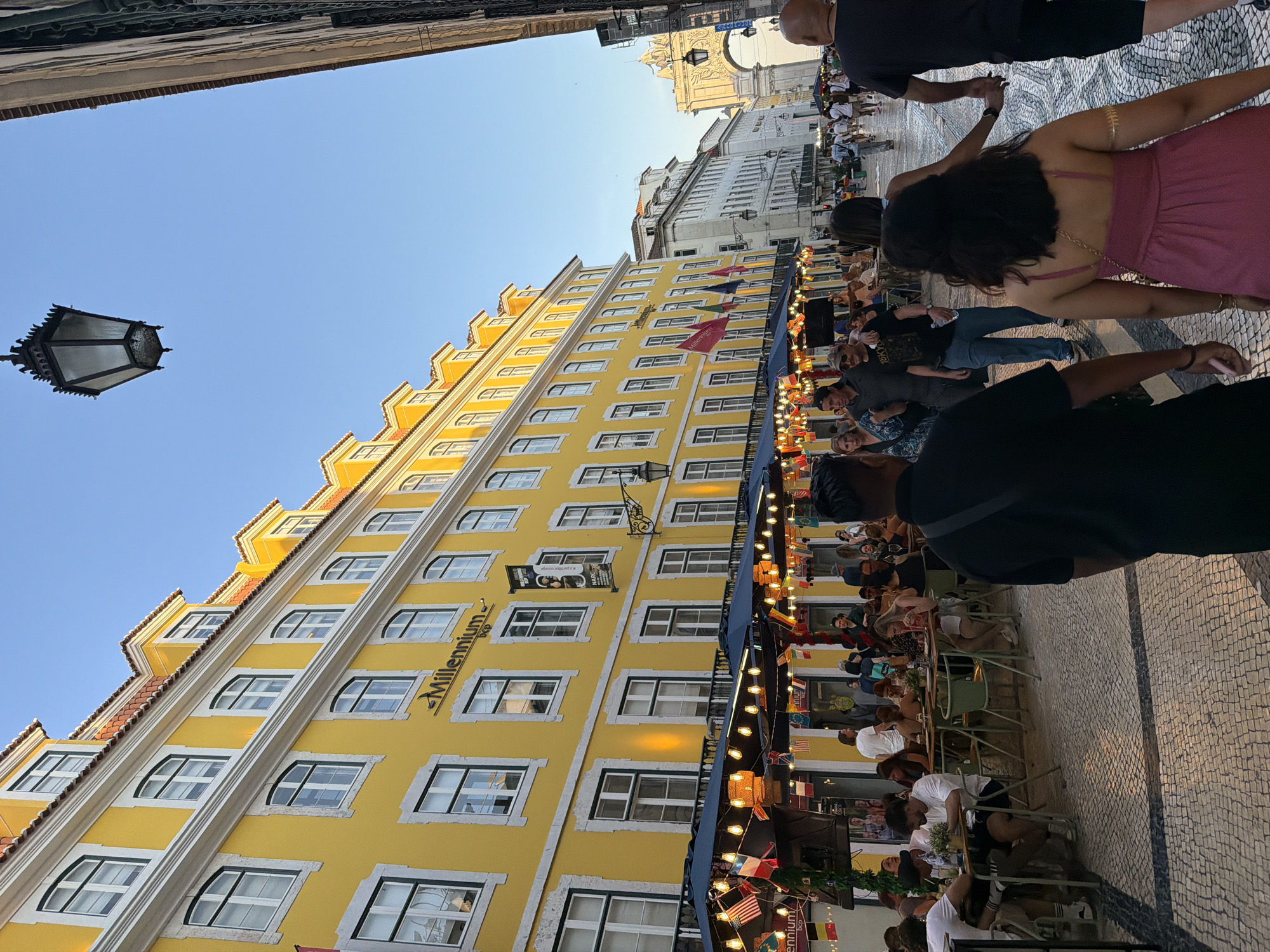The image captures a vibrant street scene in Lisbon, Portugal, likely in the Baixa district, known for its pedestrianized streets and outdoor cafes. A prominent, bright yellow building with multiple floors and arched windows dominates the left side of the frame. Flags and string lights are hung across the facade of the building, above a row of outdoor tables where people are dining.
In the foreground, several people are walking along the cobblestone street. A man in a black shirt and dark pants is walking away from the camera, while a woman in a pink dress walks in the same direction, holding hands with another person. Other individuals are visible in the background, seated at tables or walking down the street. The sky is clear and blue, suggesting it is daytime, likely late afternoon or early evening given the lighting.
The architecture is characteristic of Lisbon, with ornate details on the buildings and a classic street lamp hanging overhead. In the distance, an archway is visible, possibly the Arco da Rua Augusta. The overall atmosphere is lively and social, with a focus on outdoor dining and pedestrian activity.