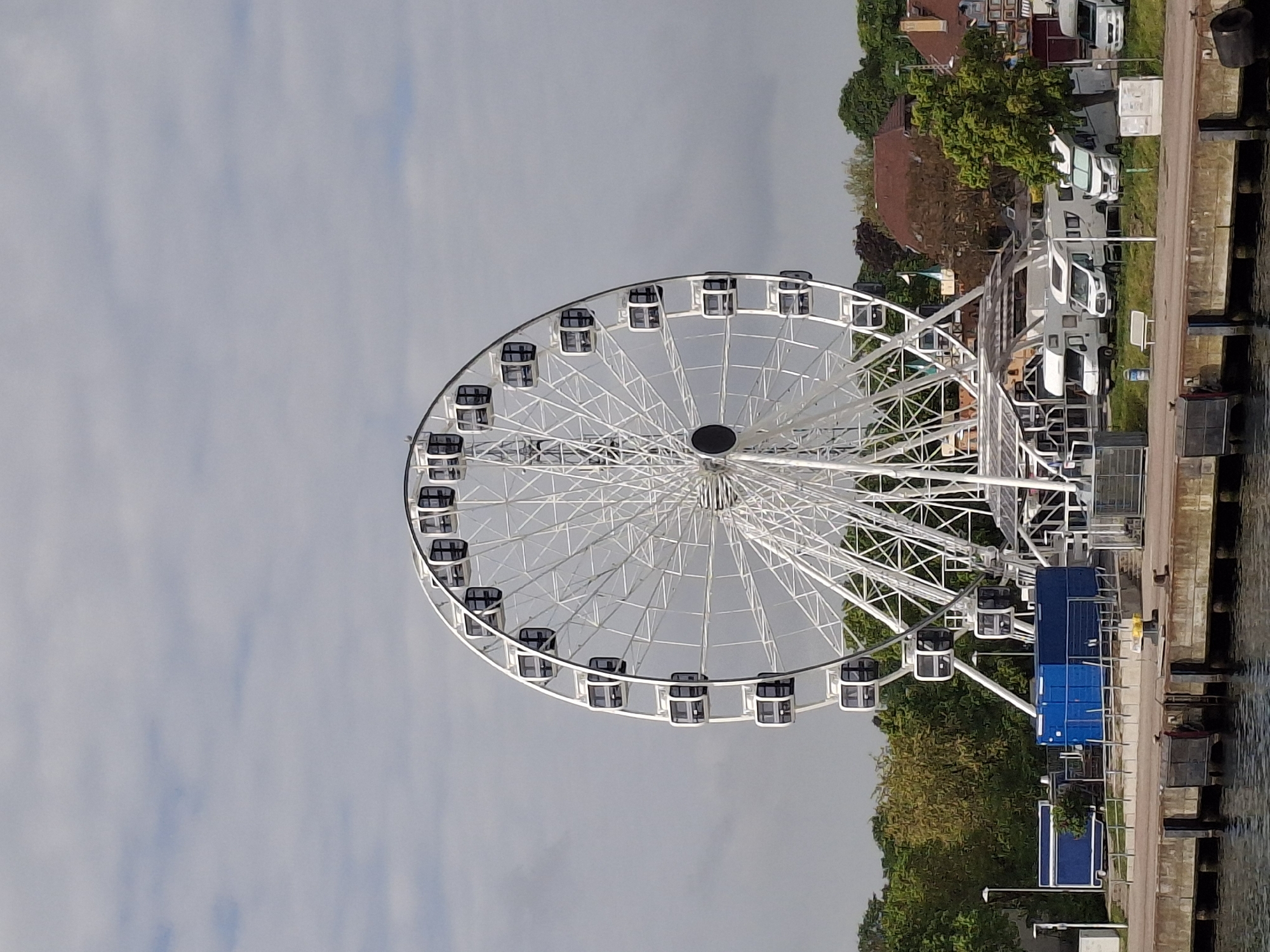 The image shows a Ferris wheel situated at the harbor in Rostock, Germany. The wheel is white with enclosed gondolas. It stands next to a waterfront promenade, bordered by a body of water on one side and trees, buildings, and parked recreational vehicles on the other. The sky is overcast with a grey hue.